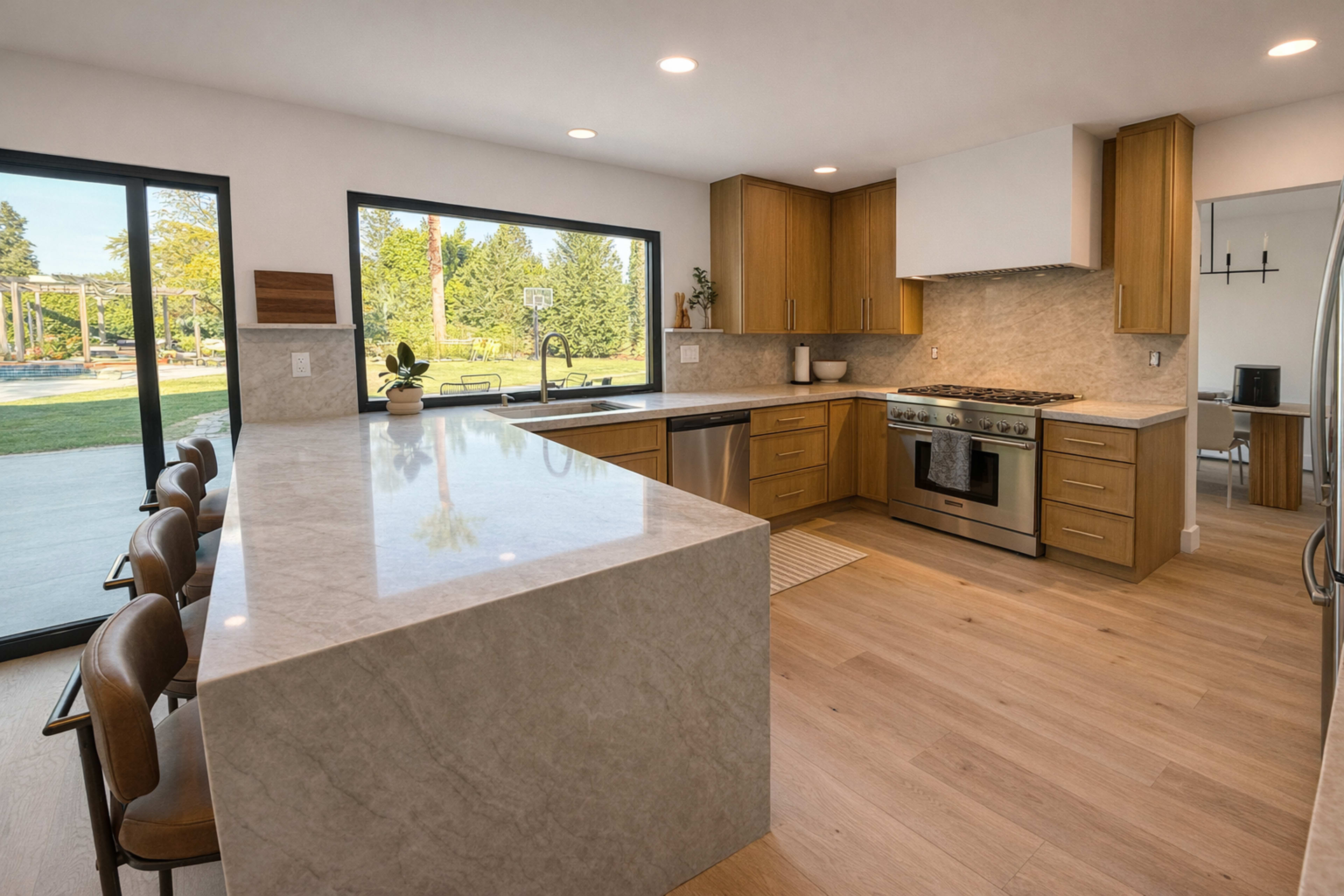 The image shows a modern kitchen with wooden cabinetry, a large island with a marble countertop, stainless steel appliances, and large windows overlooking an outdoor space.