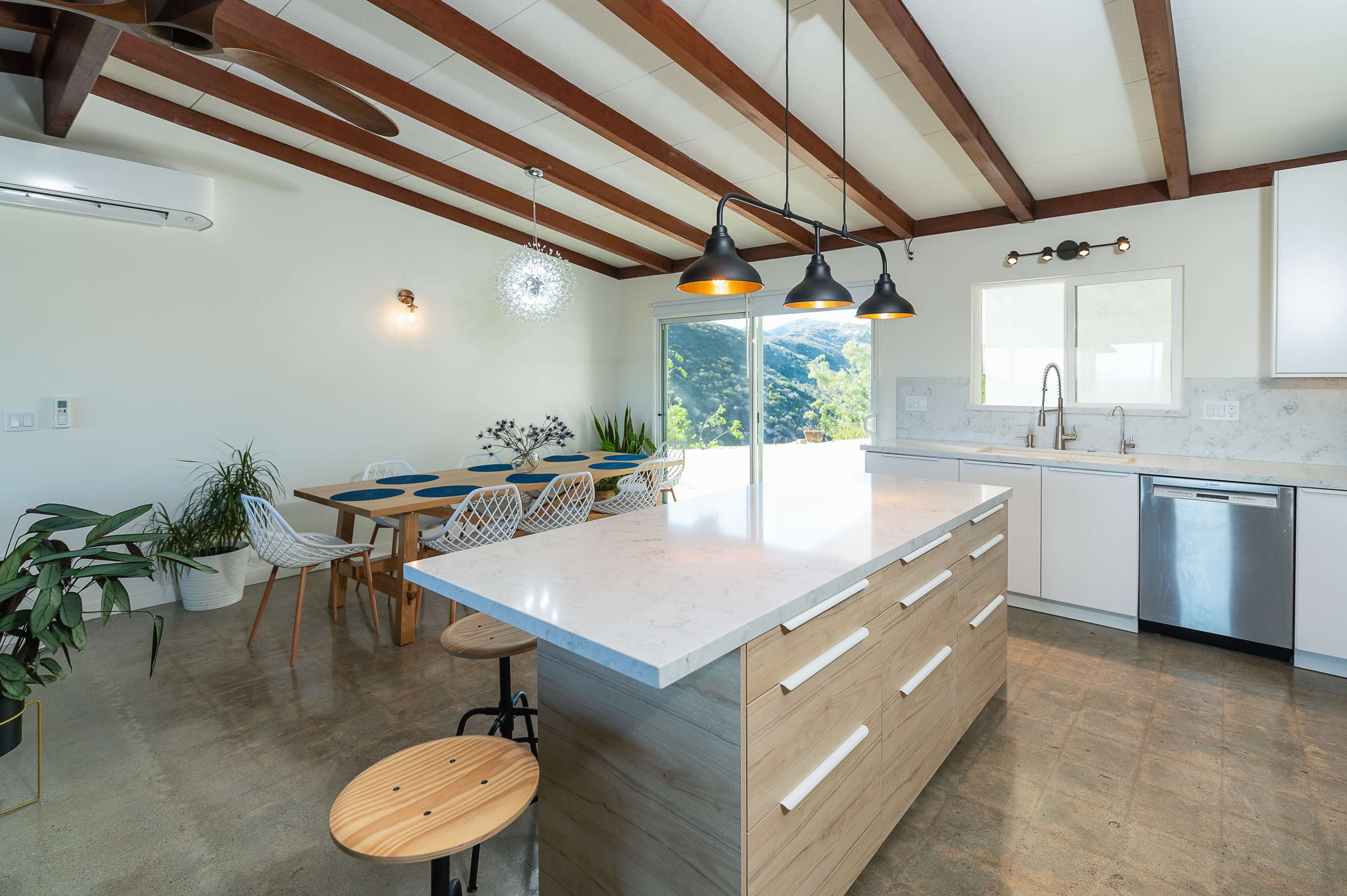 A modern kitchen features a central island with a light wood finish, pendant lighting, and a dining area with a wooden table and white chairs beside large windows showcasing a scenic view.