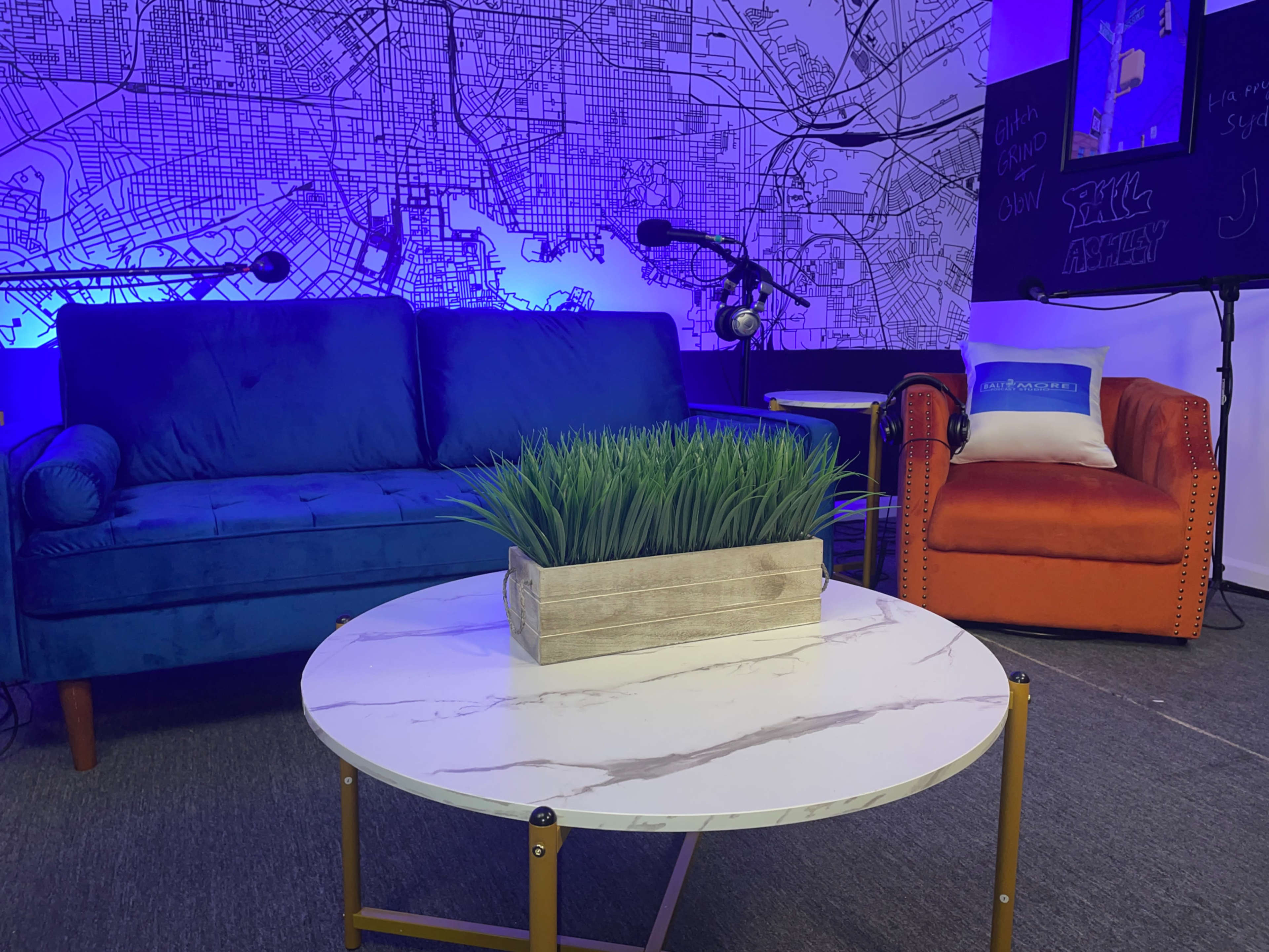 The image shows a modern lounge area with a blue couch, an orange chair, a marble coffee table, and a backdrop featuring a map design illuminated by blue lighting.