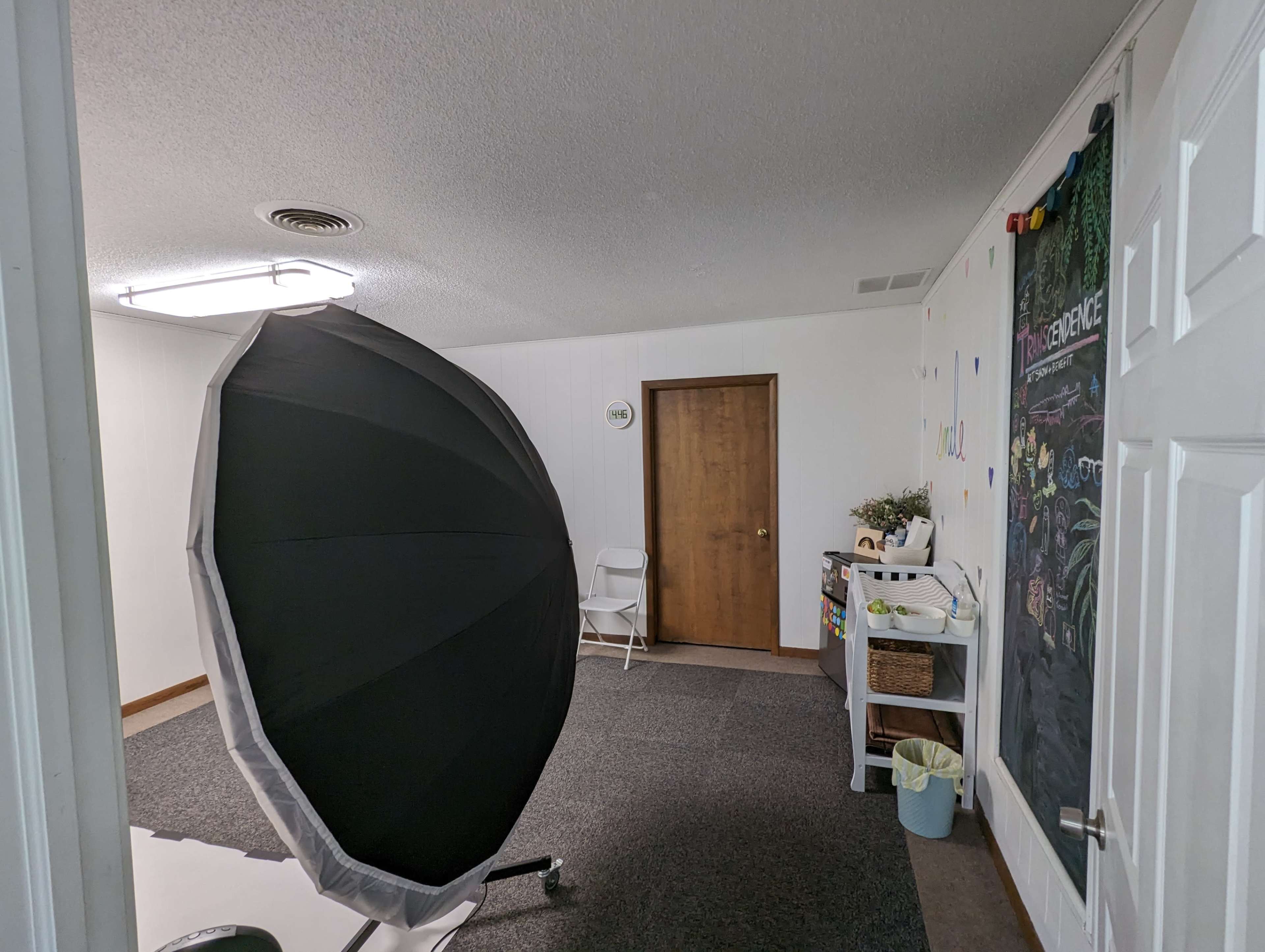A well-lit room featuring a large black umbrella-style light softbox, a closed wooden door, and a decorated wall with colorful letters.