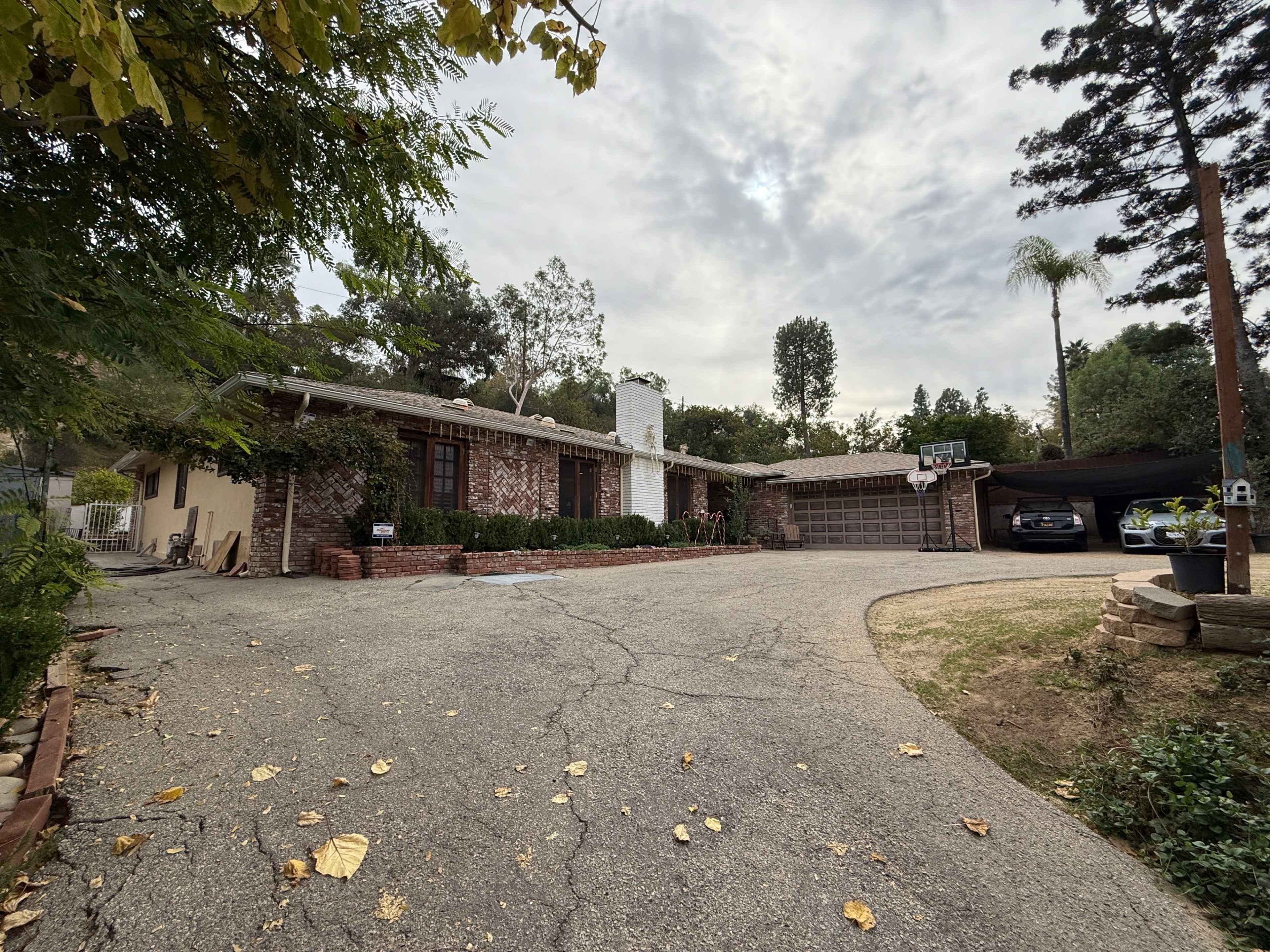 A single-story brick house with a carport is set back from a winding driveway, surrounded by greenery and under a cloudy sky.