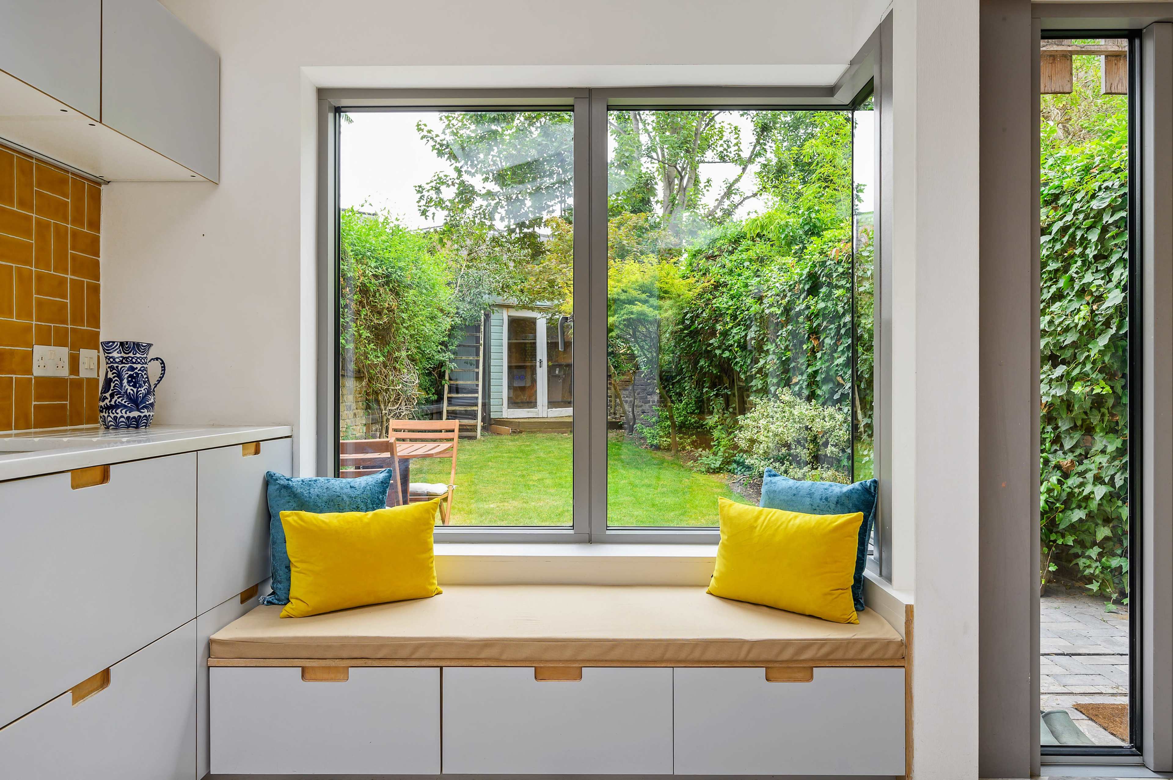 A bright kitchen nook features a cushioned bench with yellow and blue pillows, overlooking a green backyard through large windows.