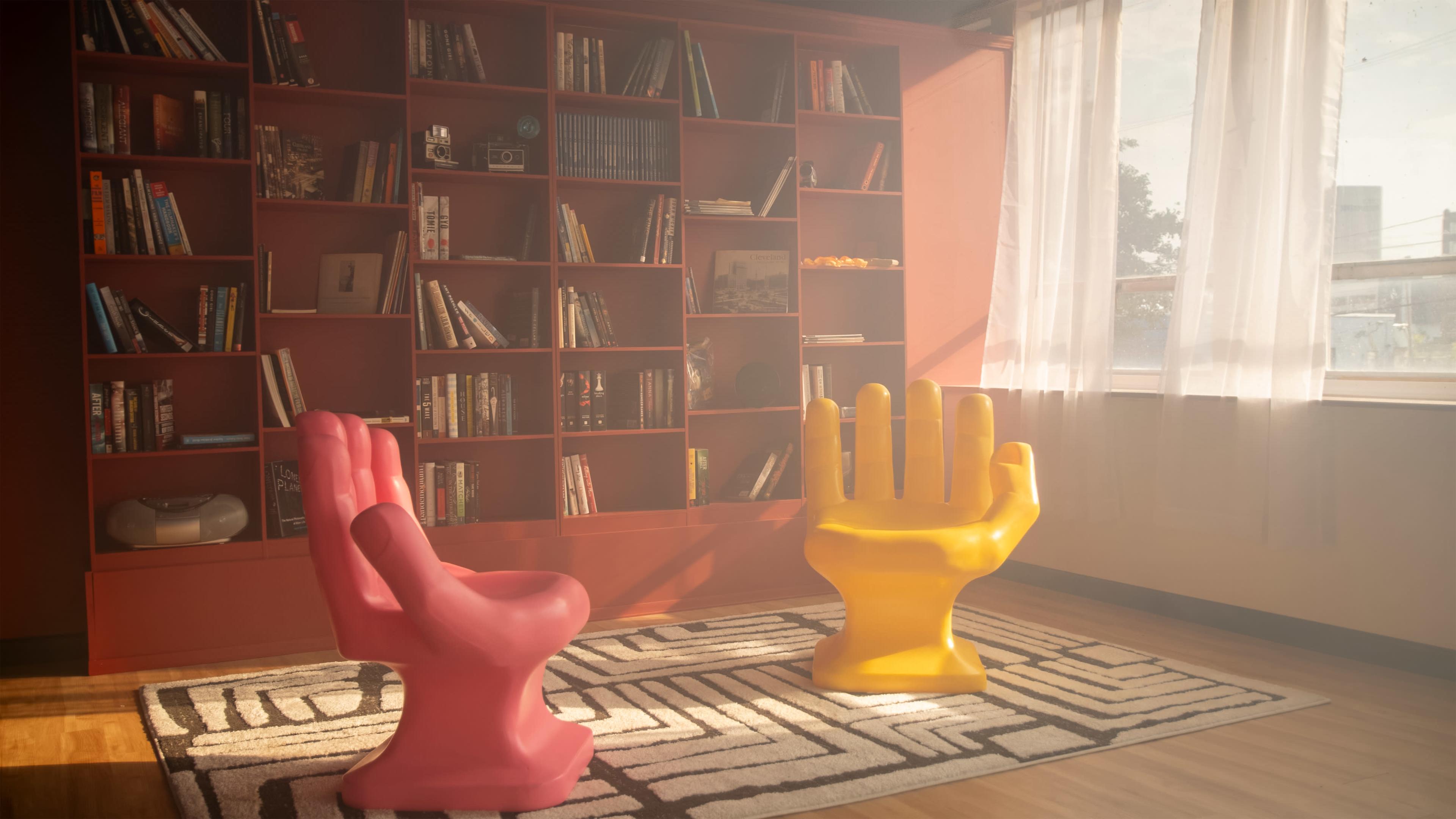 The image features a room with two colorful chair sculptures shaped like hands in pink and yellow, facing each other over a patterned rug, with a bookshelf filled with books in the background.