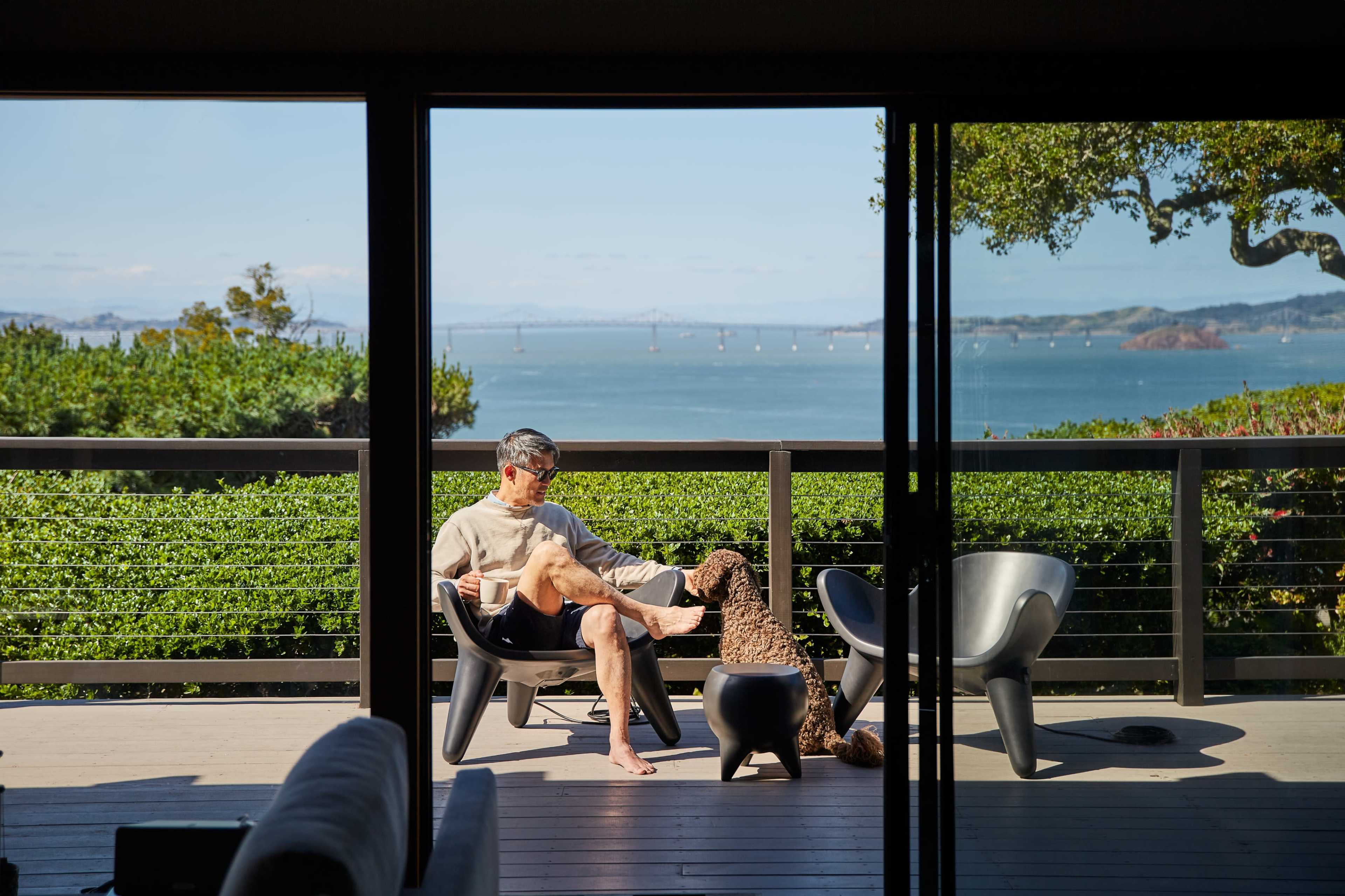 A man sits on a deck overlooking a bay while a dog approaches him.