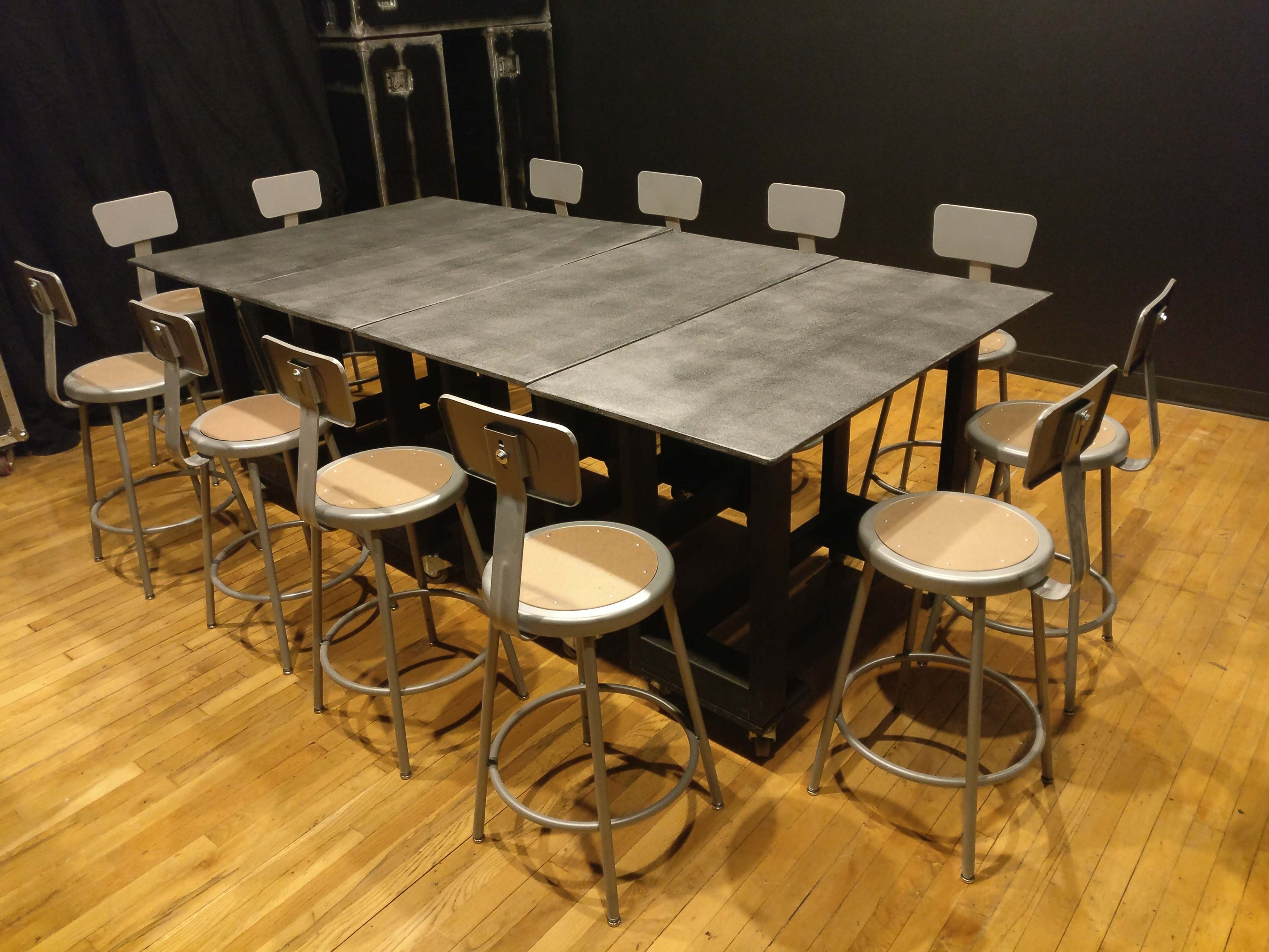 A large rectangular table with eight gray stools is set up in a room with a wooden floor and dark walls.
