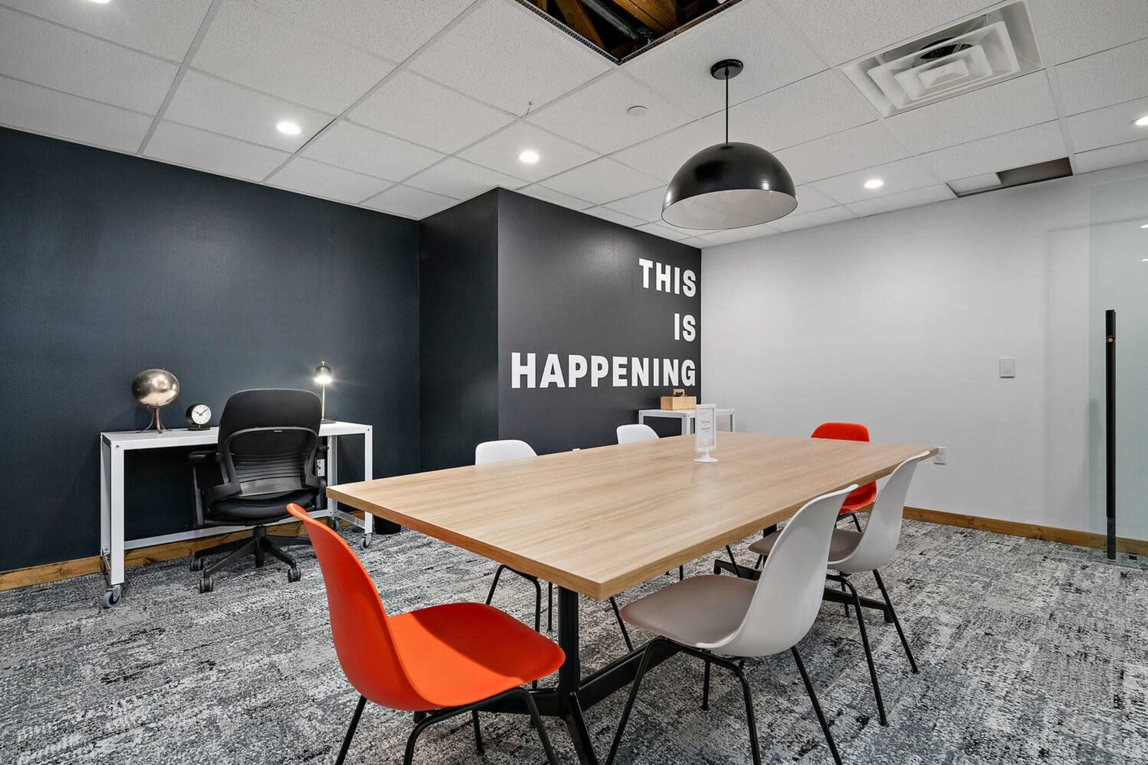 A modern meeting room features a large wooden table surrounded by colorful chairs, with a black wall displaying the text "THIS IS HAPPENING."