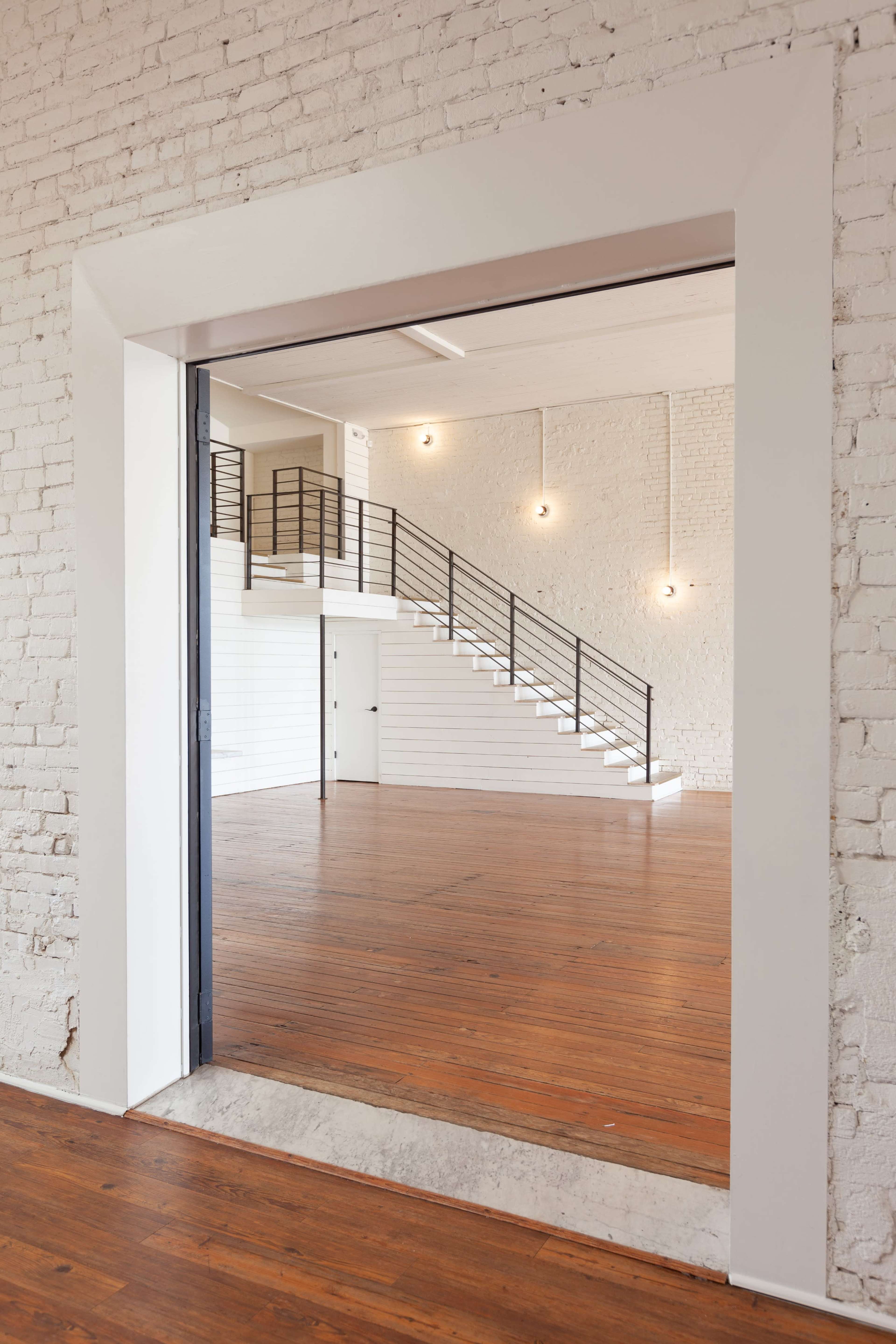 A well-lit, spacious room features a wooden floor, white brick walls, and a staircase with a black railing.