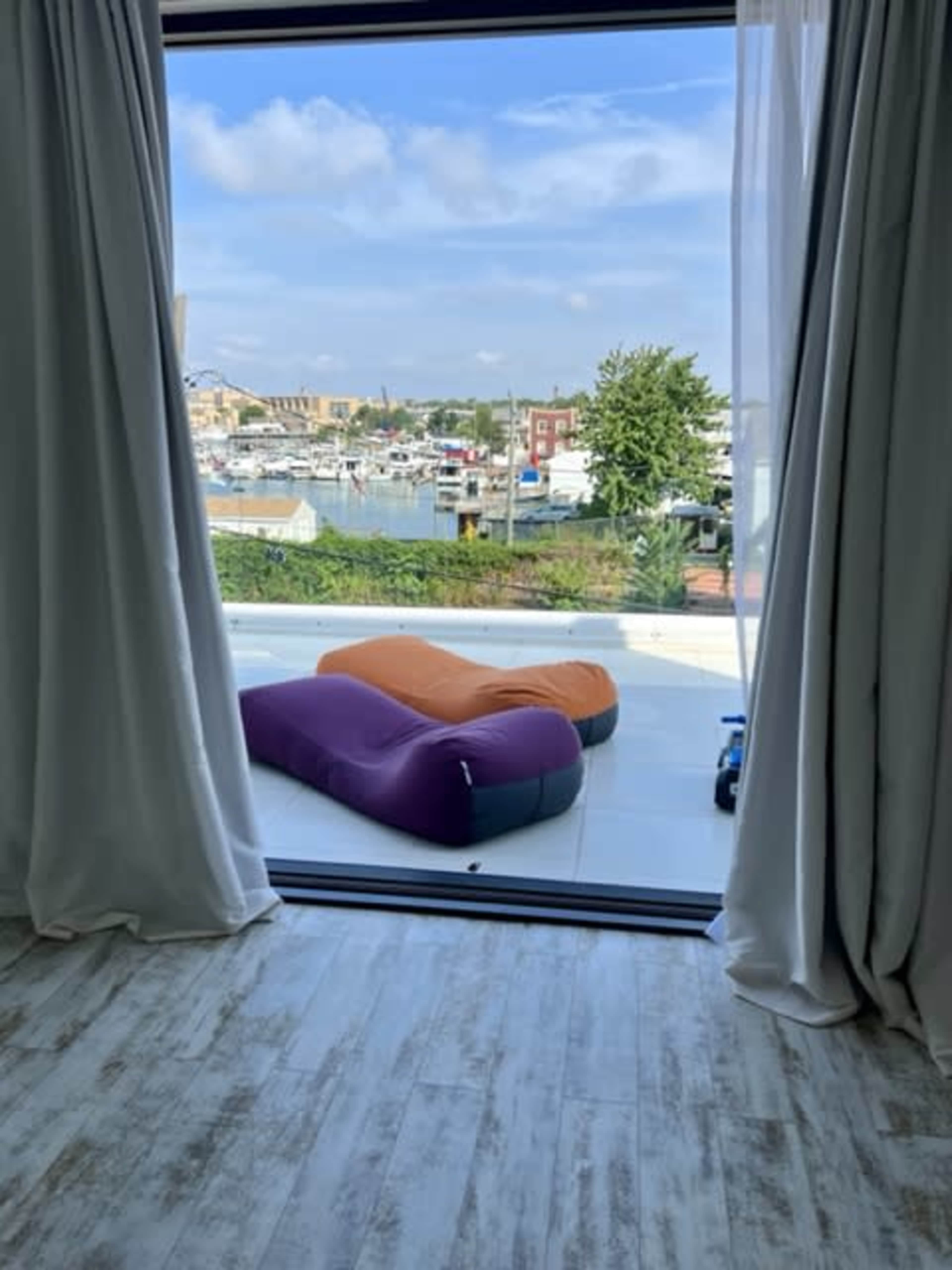 The image shows a view through a large window featuring two inflatable loungers—one purple and one orange—overlooking a marina with boats.
