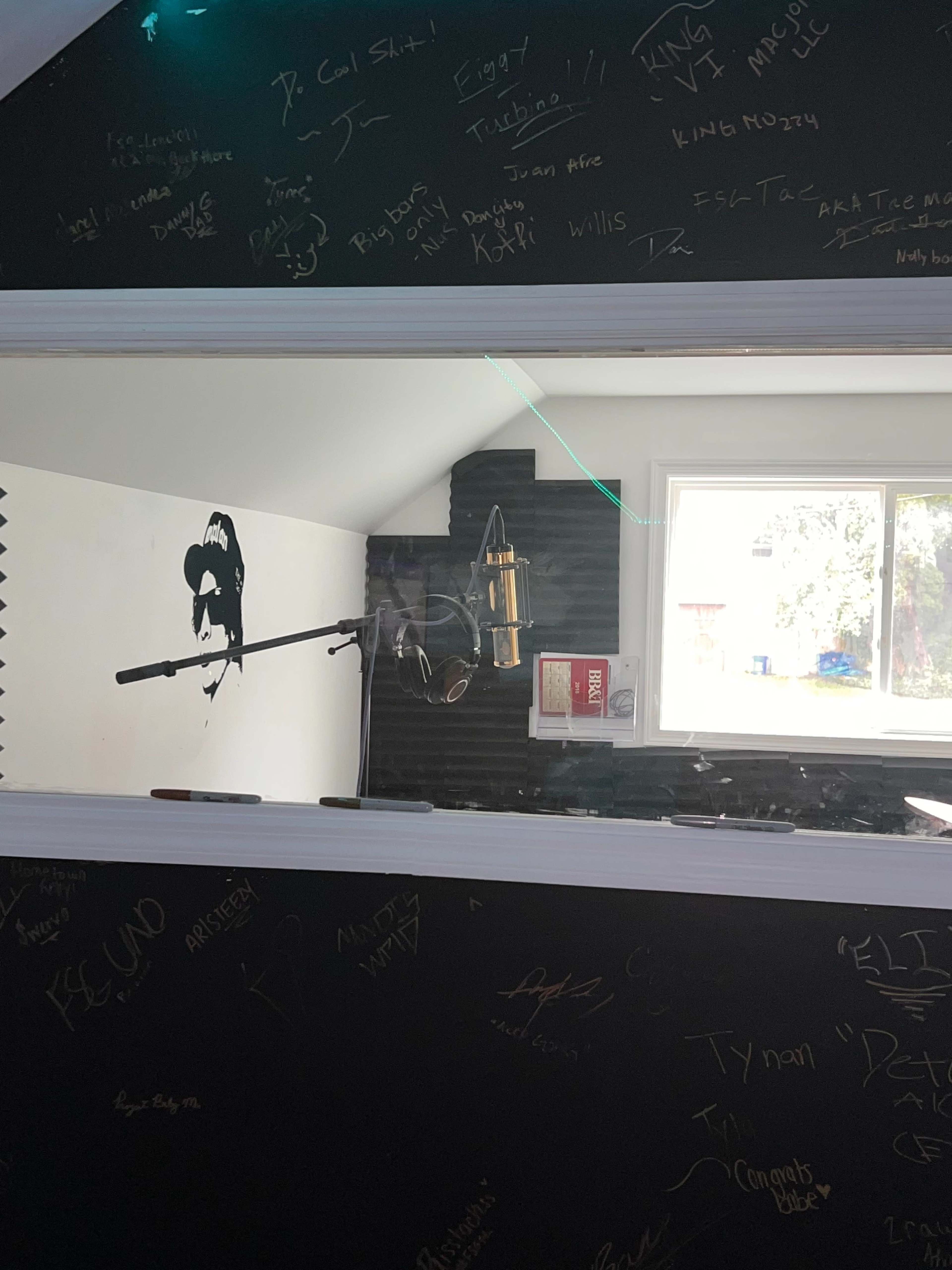 A recording studio with a microphone in front of a window displaying a wall covered in signatures and a black-and-white portrait of a person.