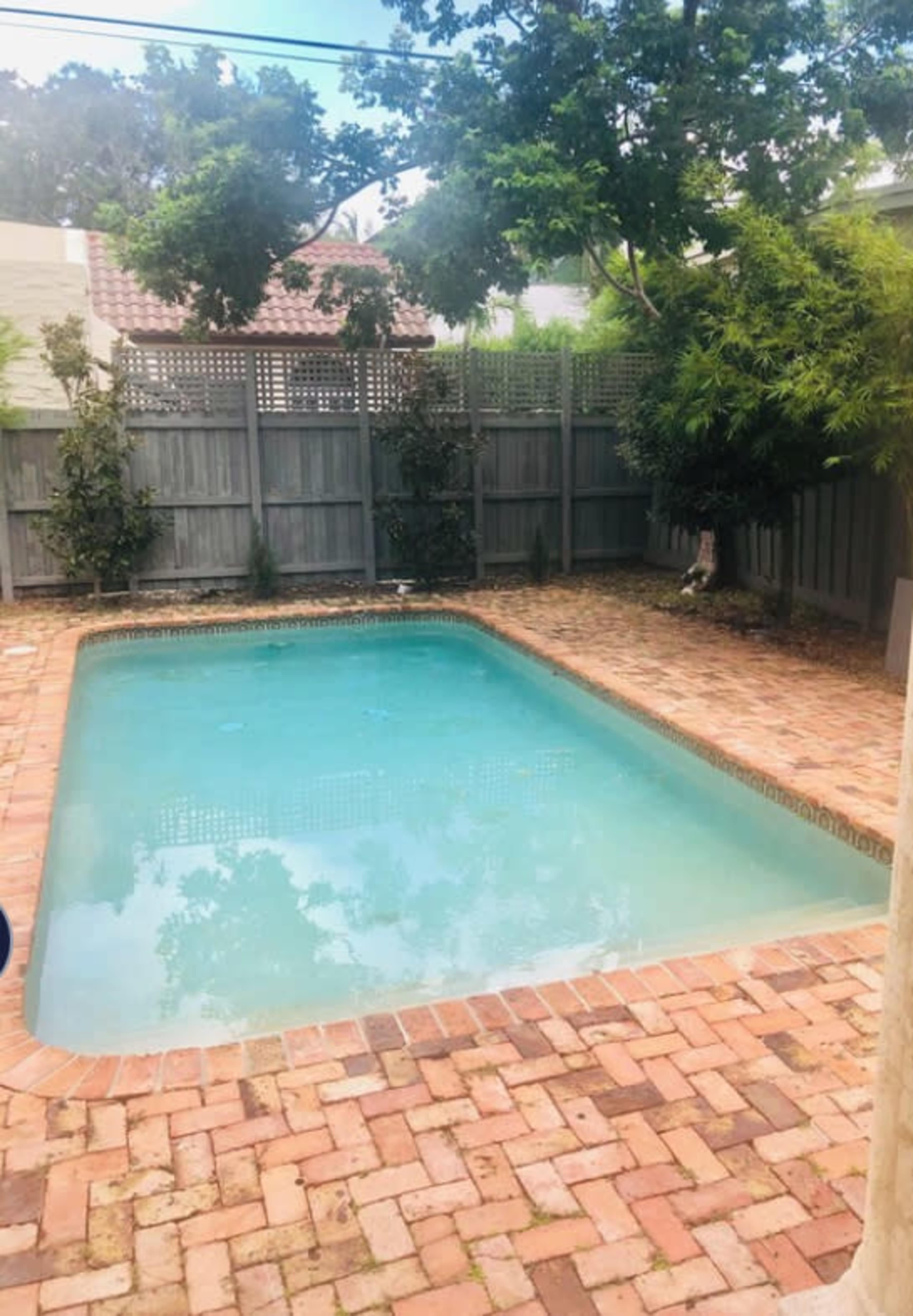 The image shows a rectangular swimming pool surrounded by a brick patio and enclosed by wooden fencing and greenery.