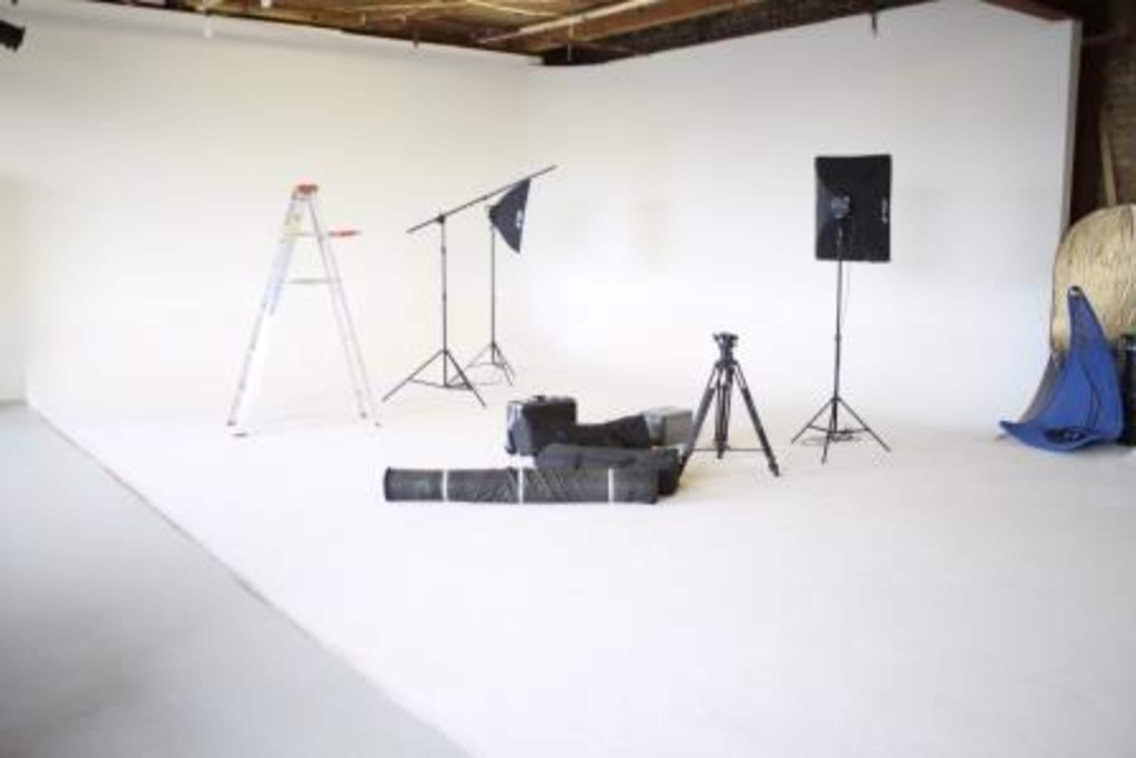 A photography studio with a ladder, lighting equipment, a tripod, and a rolled-up backdrop placed on a plain white floor.