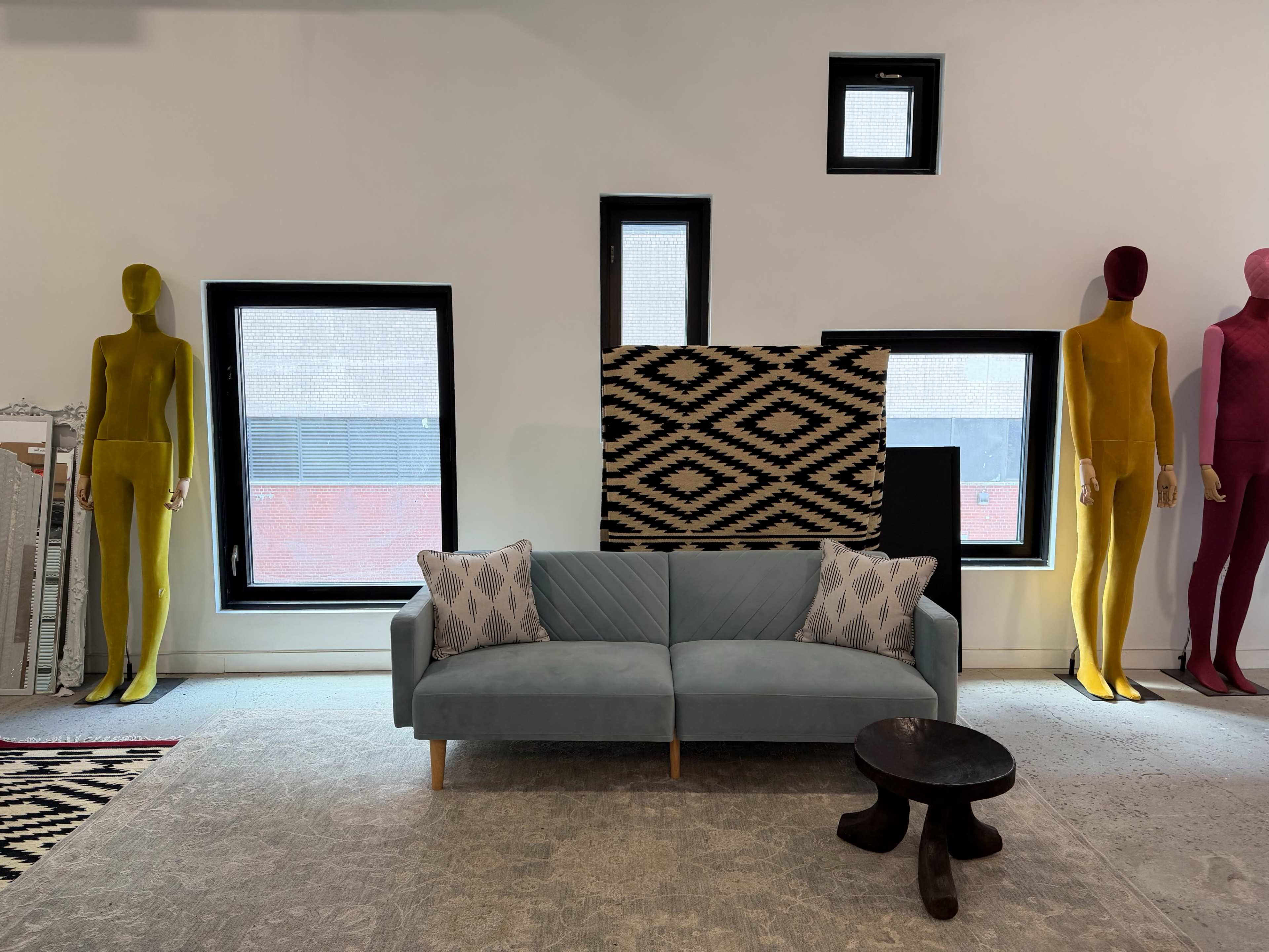 The image shows a modern living space featuring a blue sofa with decorative pillows, flanked by two colorful mannequin figures and a patterned wall hanging against a backdrop of large windows.
