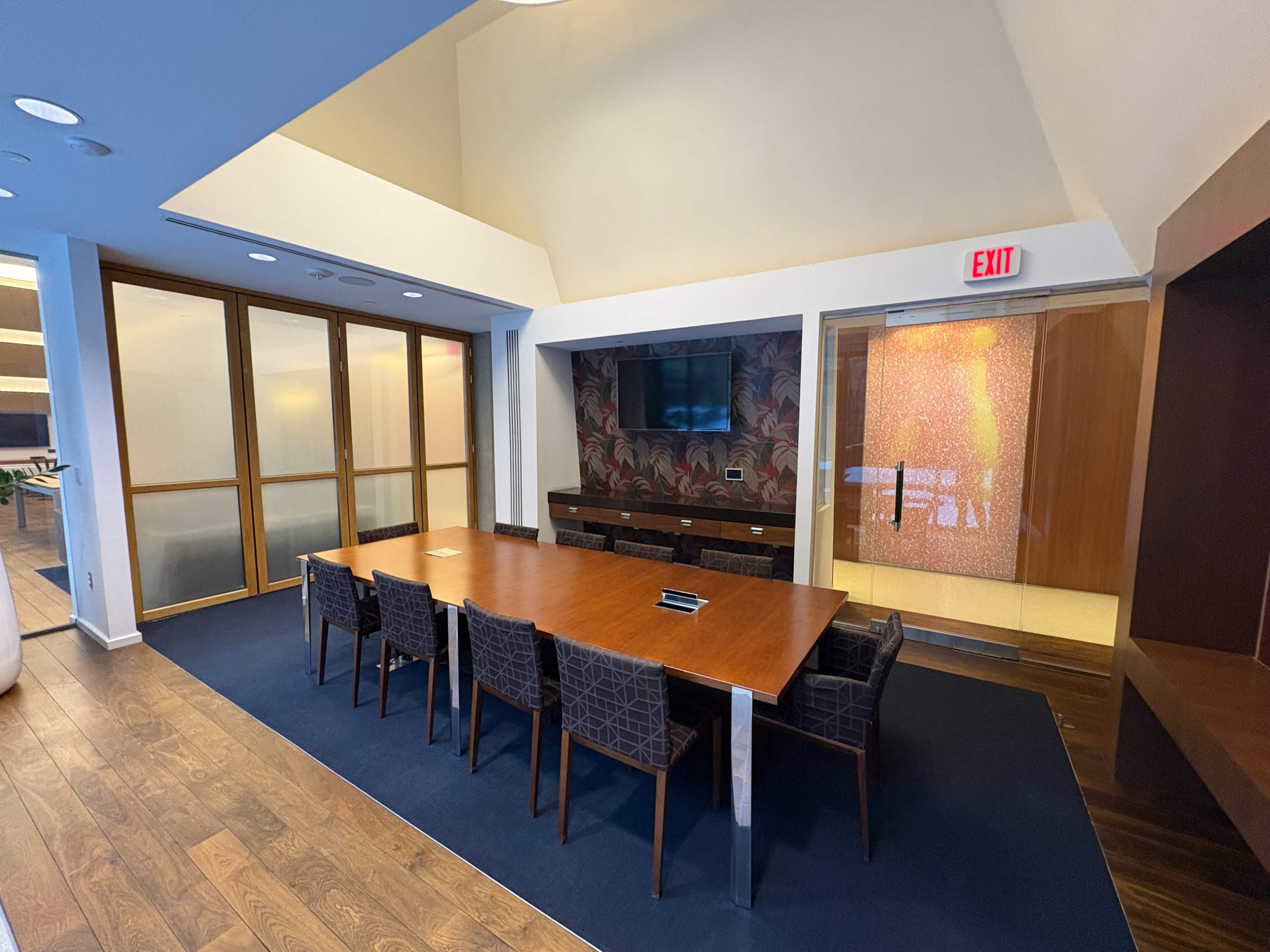 The image shows a modern conference room featuring a large wooden table surrounded by dark upholstered chairs, with a wall-mounted screen and glass doors leading to another space.