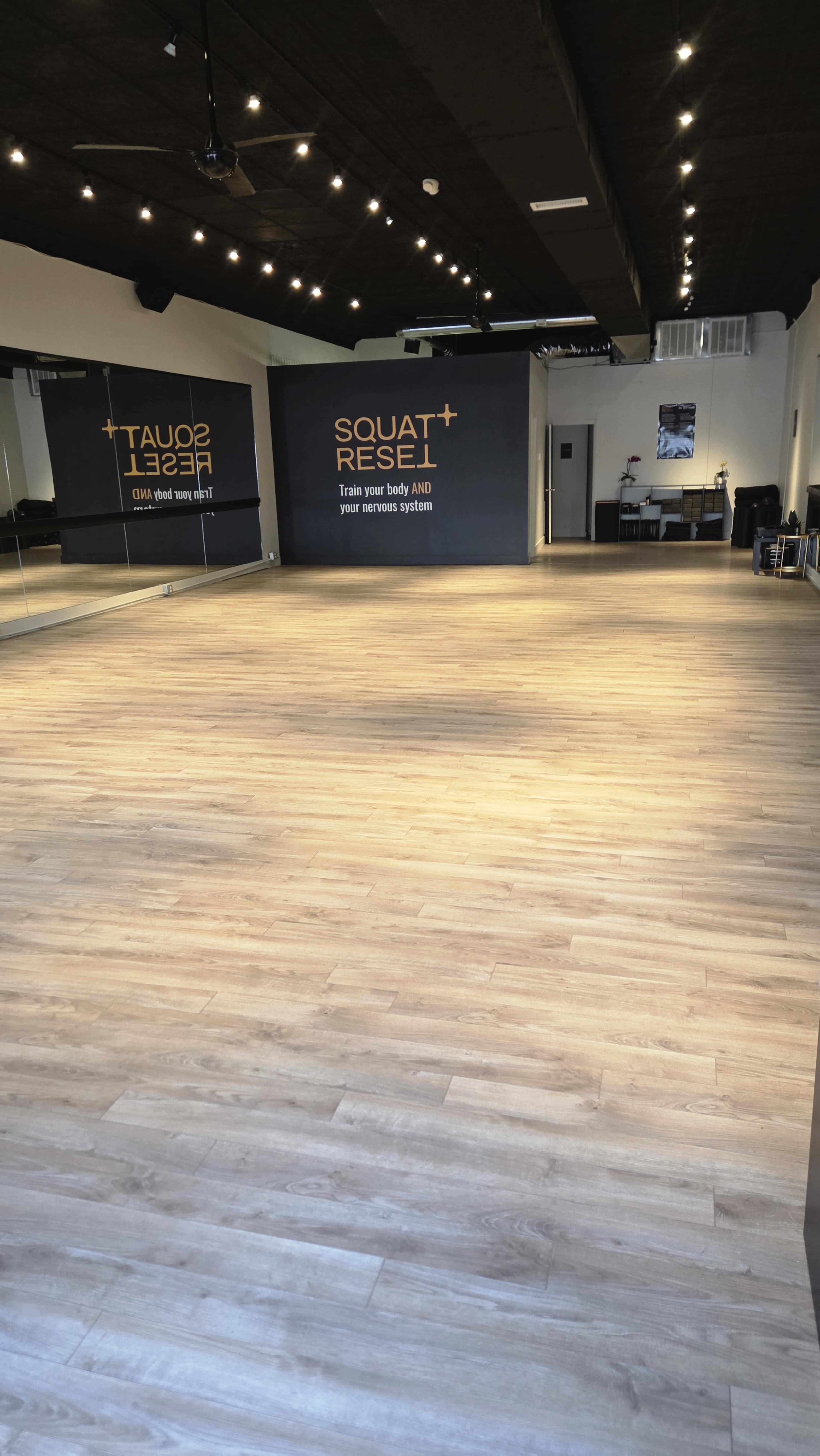 The image shows a bright, spacious fitness studio with wooden flooring, large mirrors on one wall, and a prominent wall sign that reads "SQUAT + RESET."
