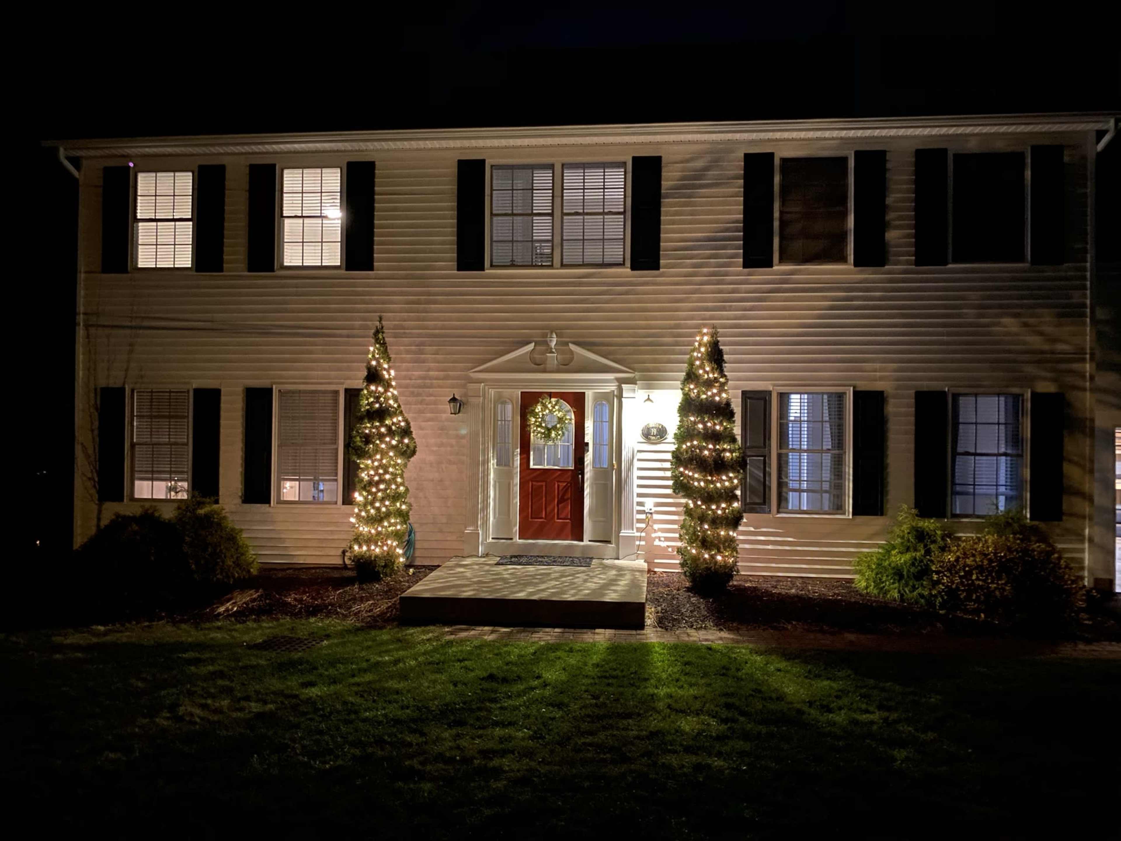 A two-story house is decorated with holiday lights on either side of the front entrance, featuring a wreath on the door and well-lit conifer trees.
