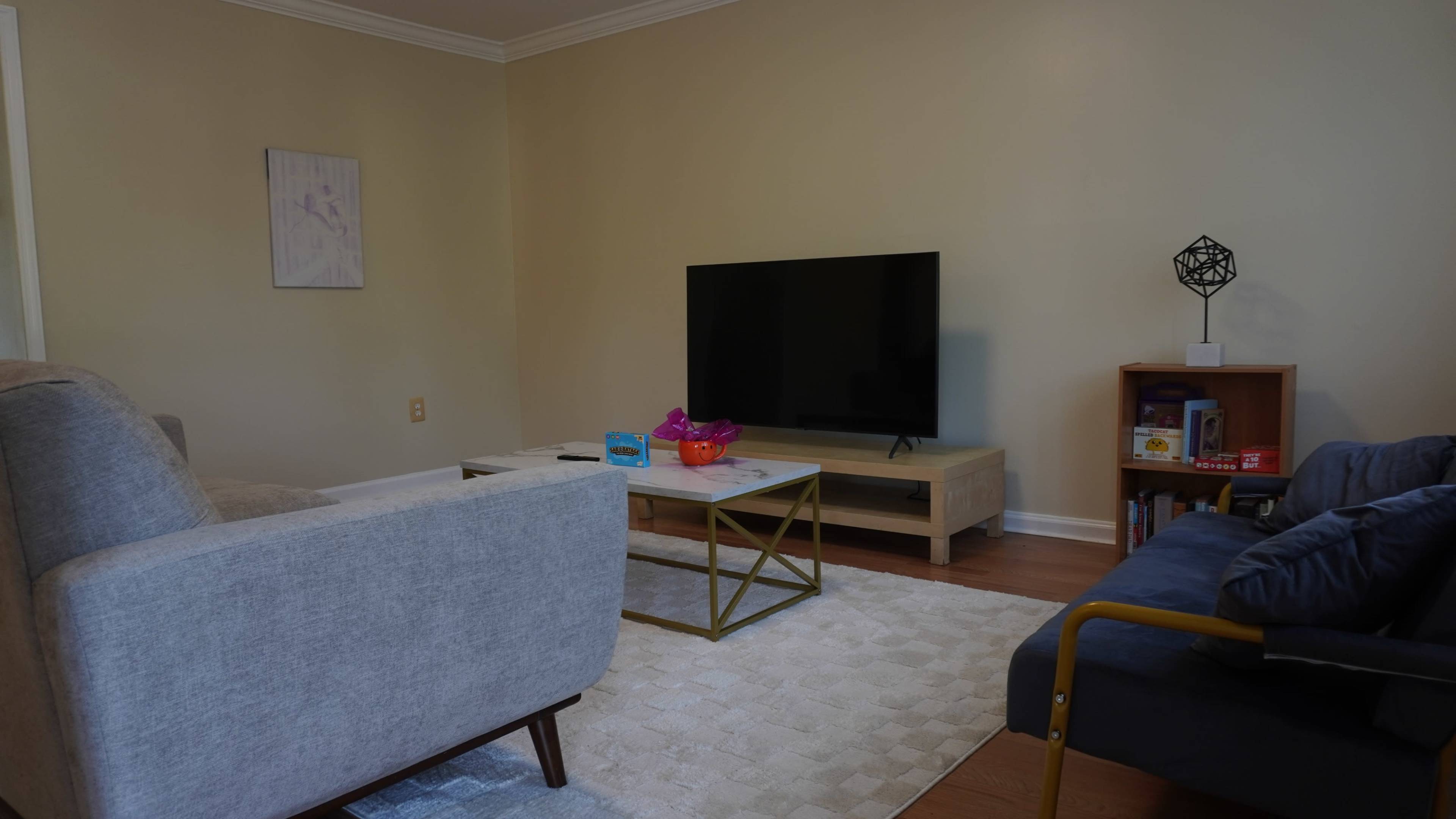 A living room with a gray sofa, a blue armchair, a coffee table, a large television on a wooden stand, and a small bookshelf against the wall.