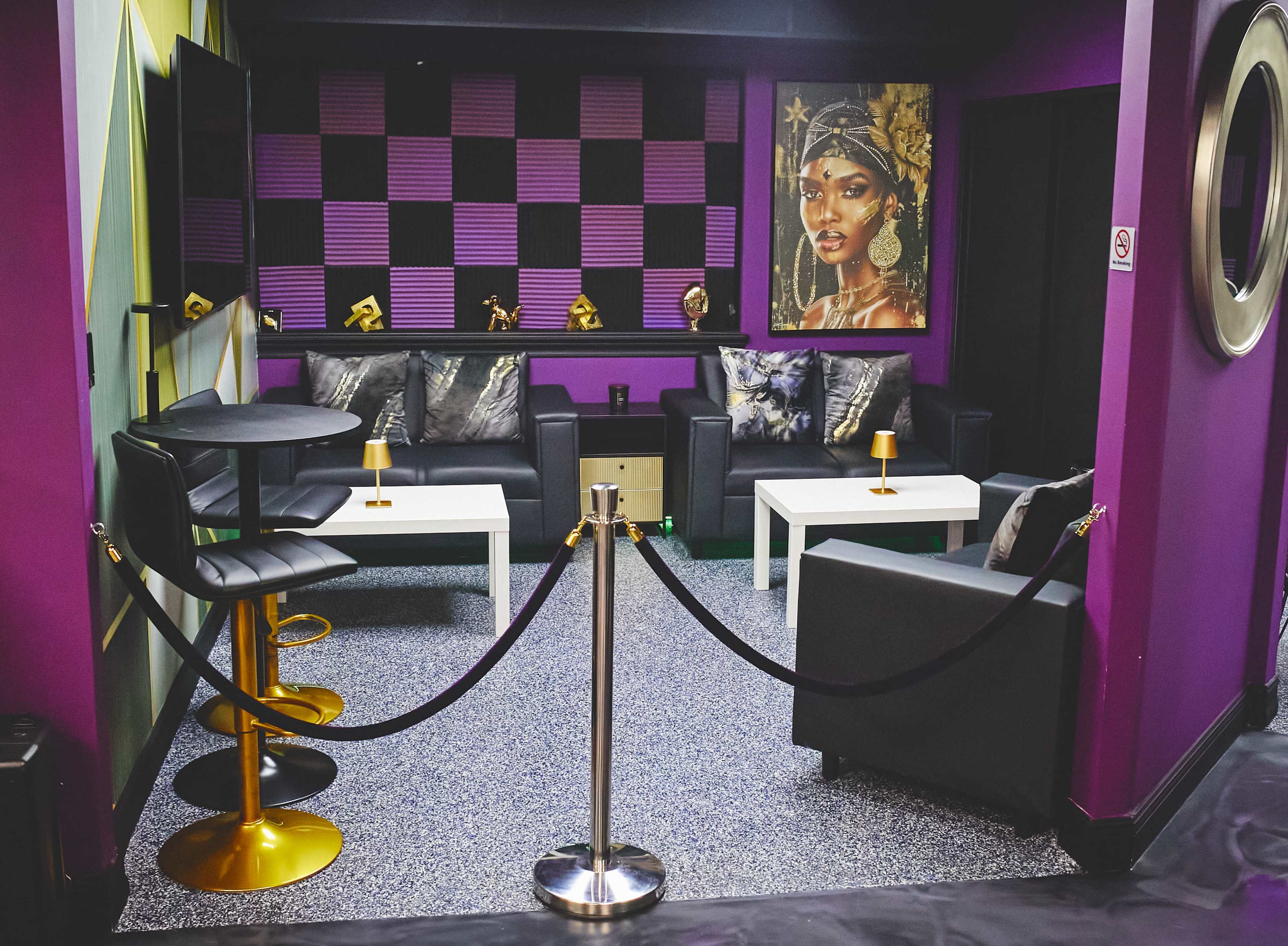 The image shows a vibrant lounge area with black and purple decor, featuring two black leather sofas, white tables, and a large portrait of a woman on the wall.