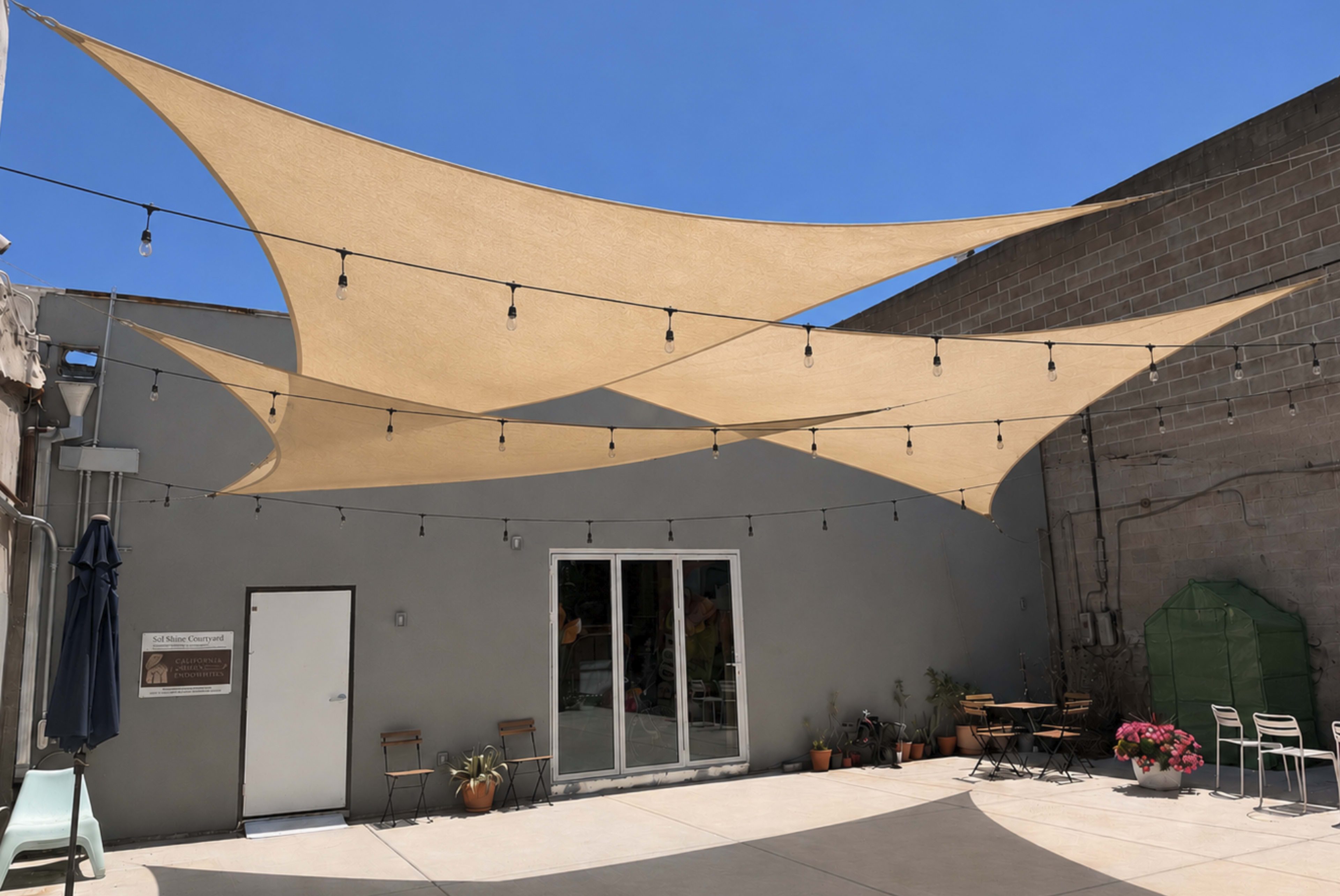 The image shows a sunlit outdoor space with large beige shade sails overhead, string lights, and potted plants arranged near a gray wall.