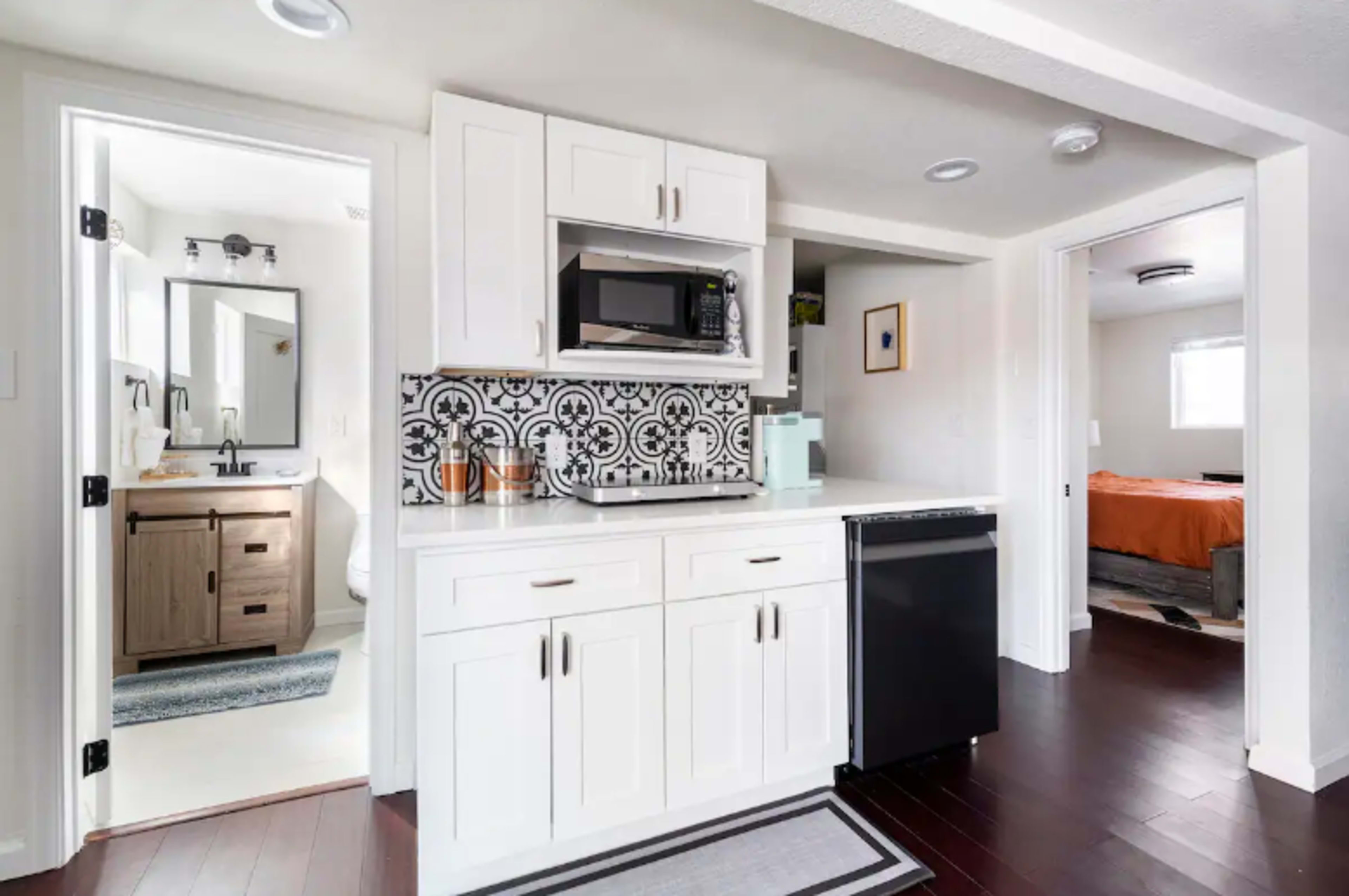 A modern kitchen with white cabinets, a microwave, and a black refrigerator, adjacent to a bathroom with a sink and a doorway leading to a bedroom.