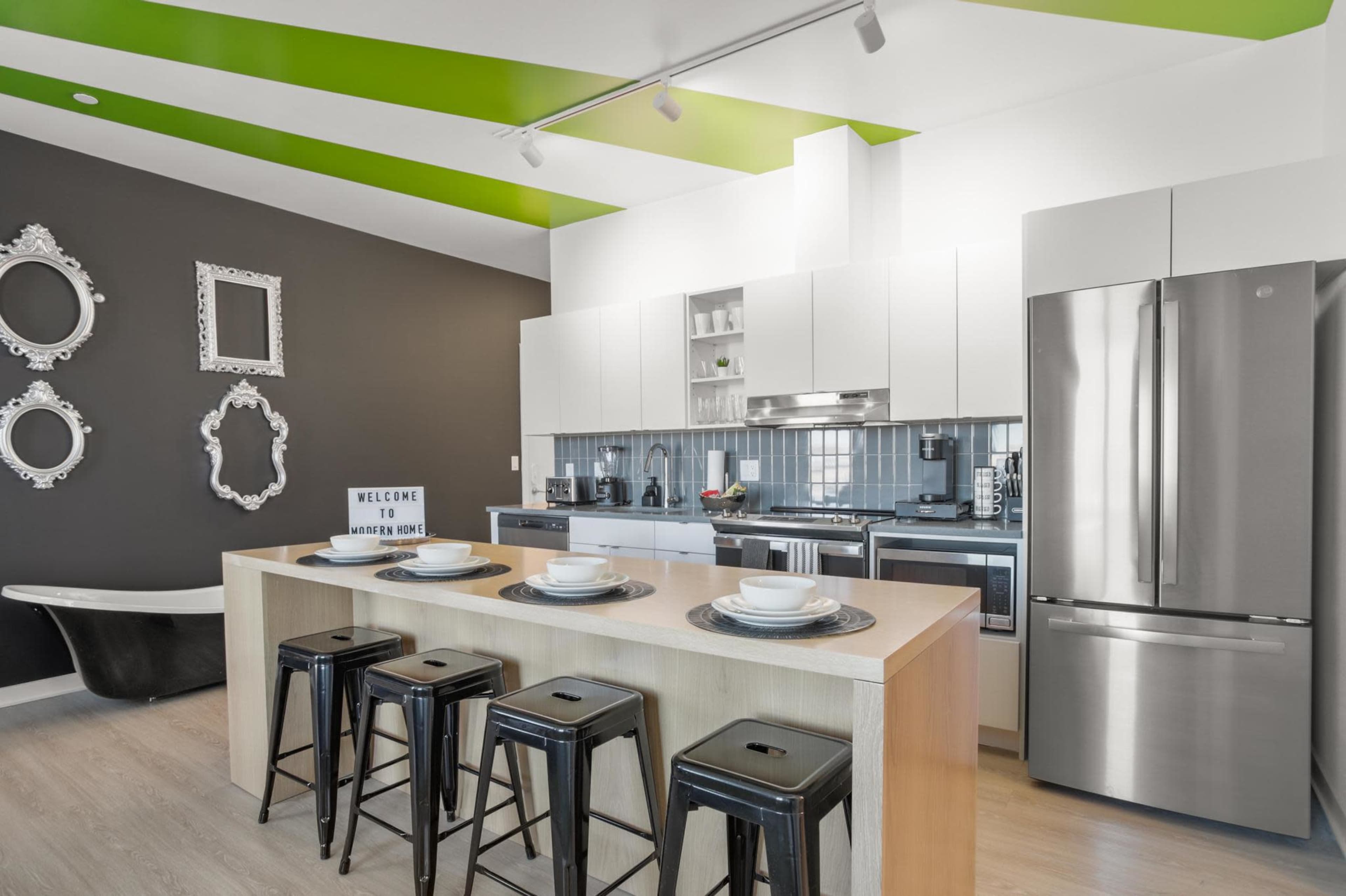 The image depicts a modern kitchen with white cabinetry, stainless steel appliances, a central island with four black stools, and a contrasting dark wall with decorative mirrors.
