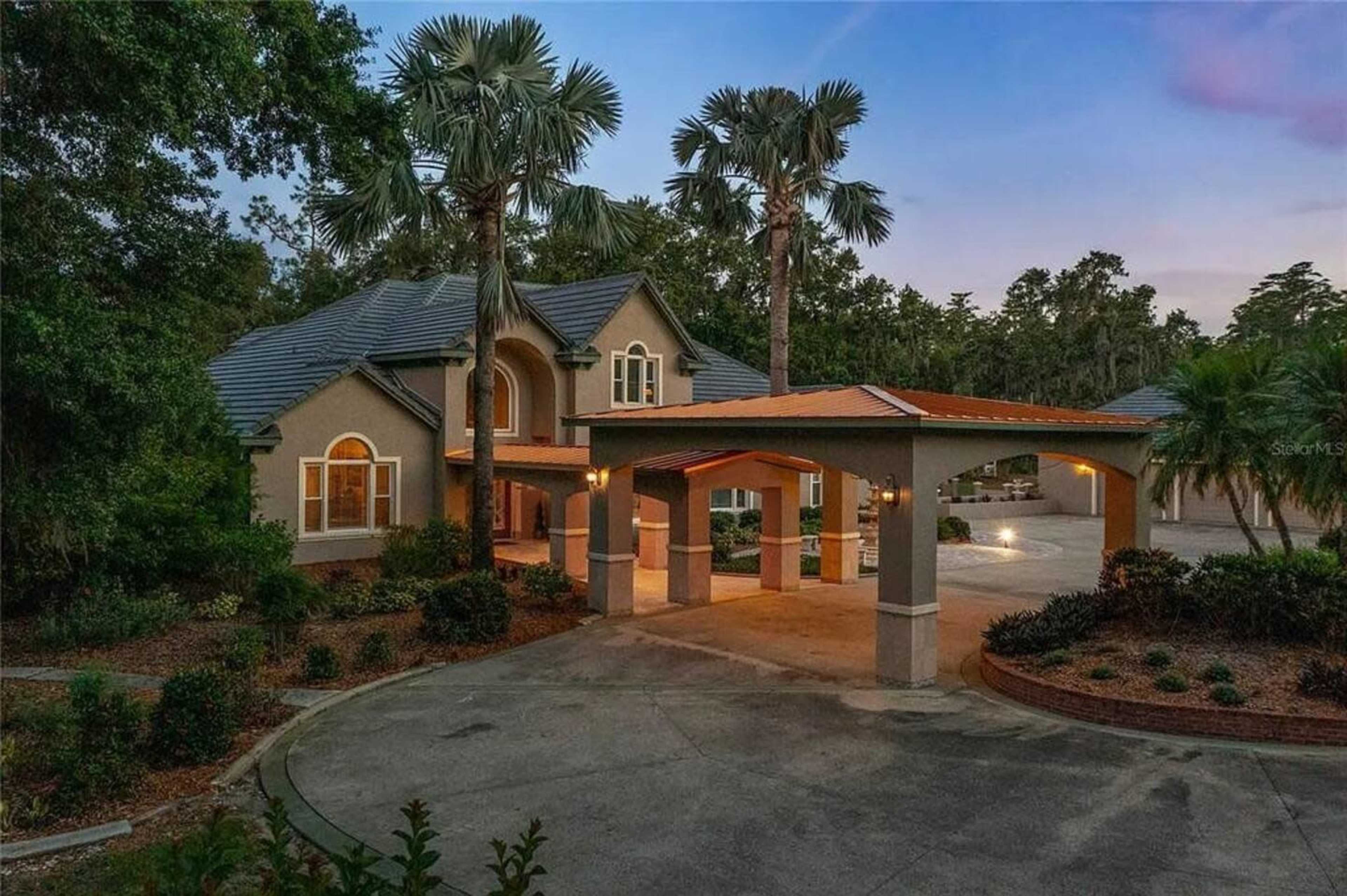 The image shows a large, two-story house with a curved driveway, palm trees, and a covered entrance.