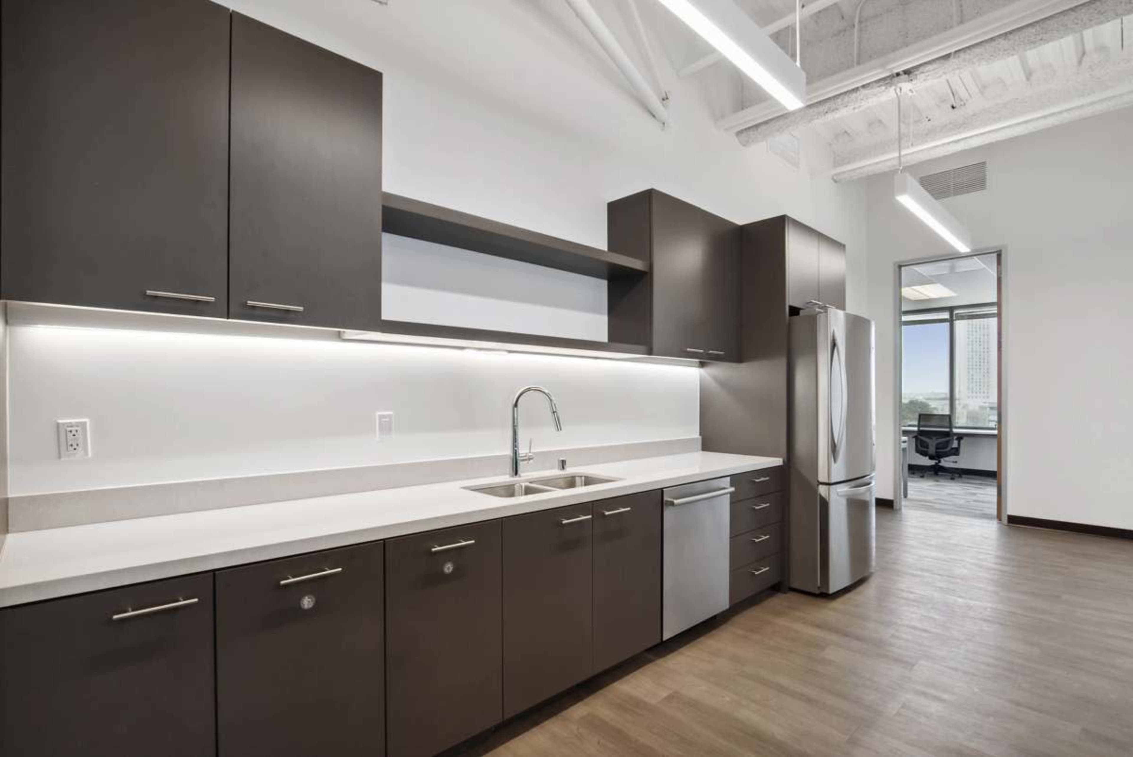 A modern kitchen with dark cabinetry, a stainless steel refrigerator, and a sink set against a light countertop in an office space.