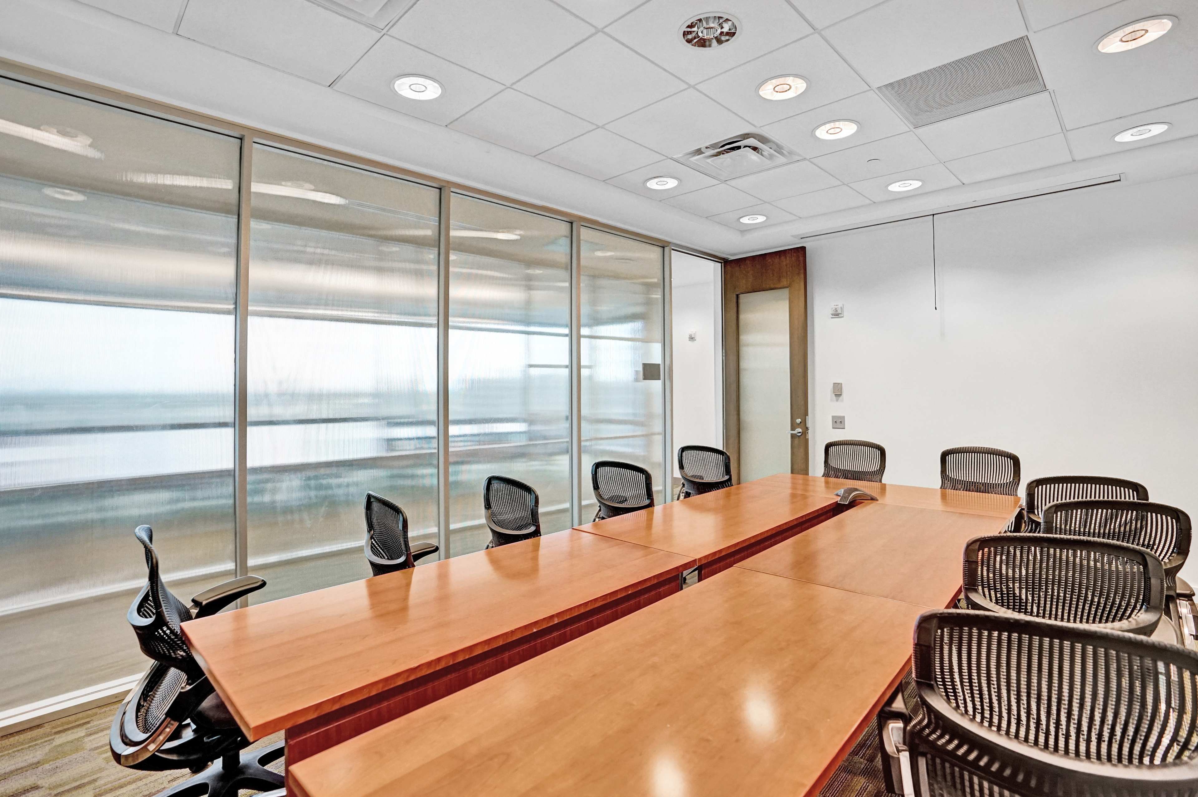 A modern conference room features a large wooden table surrounded by eight black mesh chairs, with floor-to-ceiling windows providing a view outside.