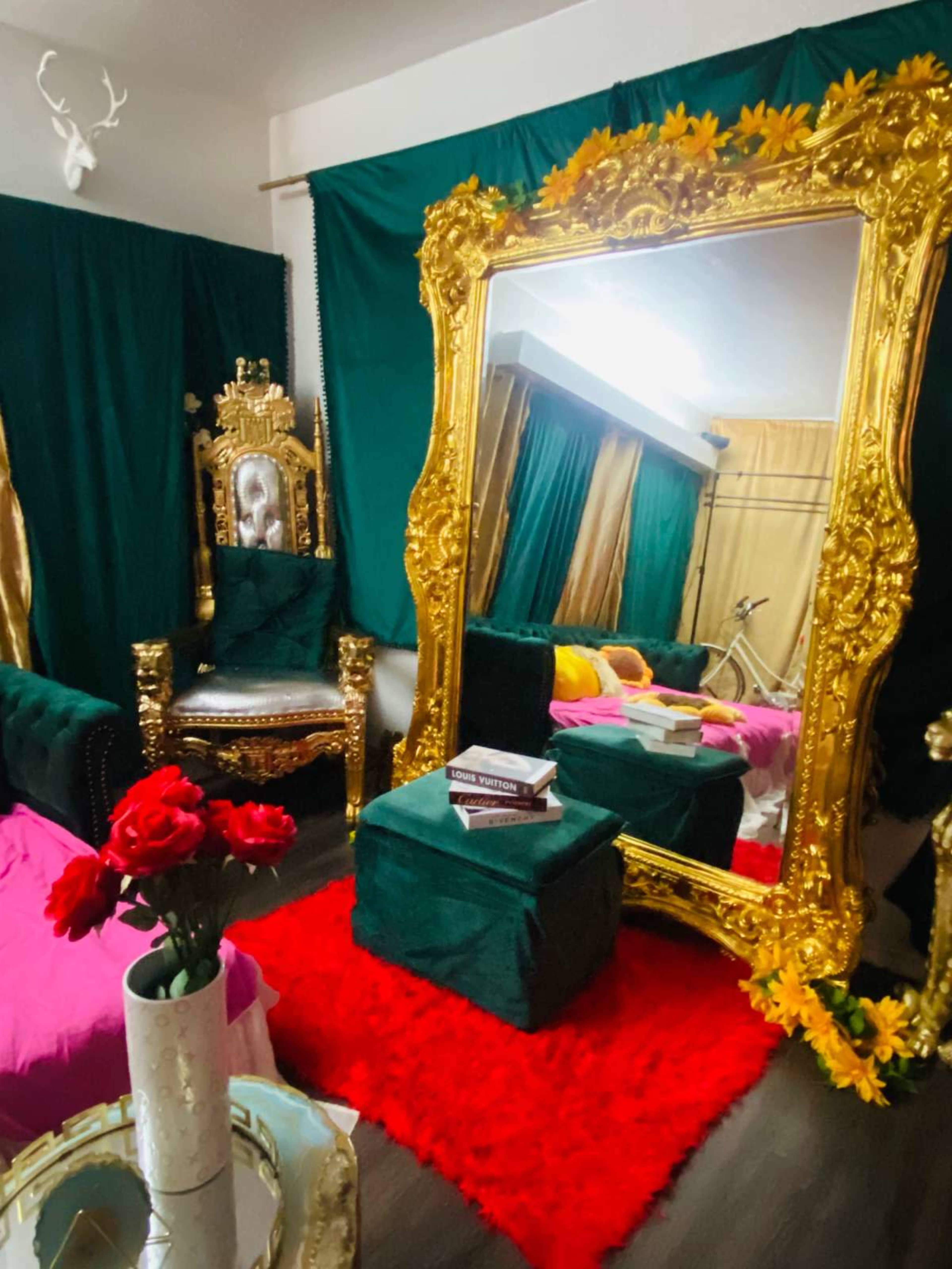 The room features a large ornate mirror, a golden throne-like chair, a pink seating area, and a vibrant red rug adorned with a vase of roses.