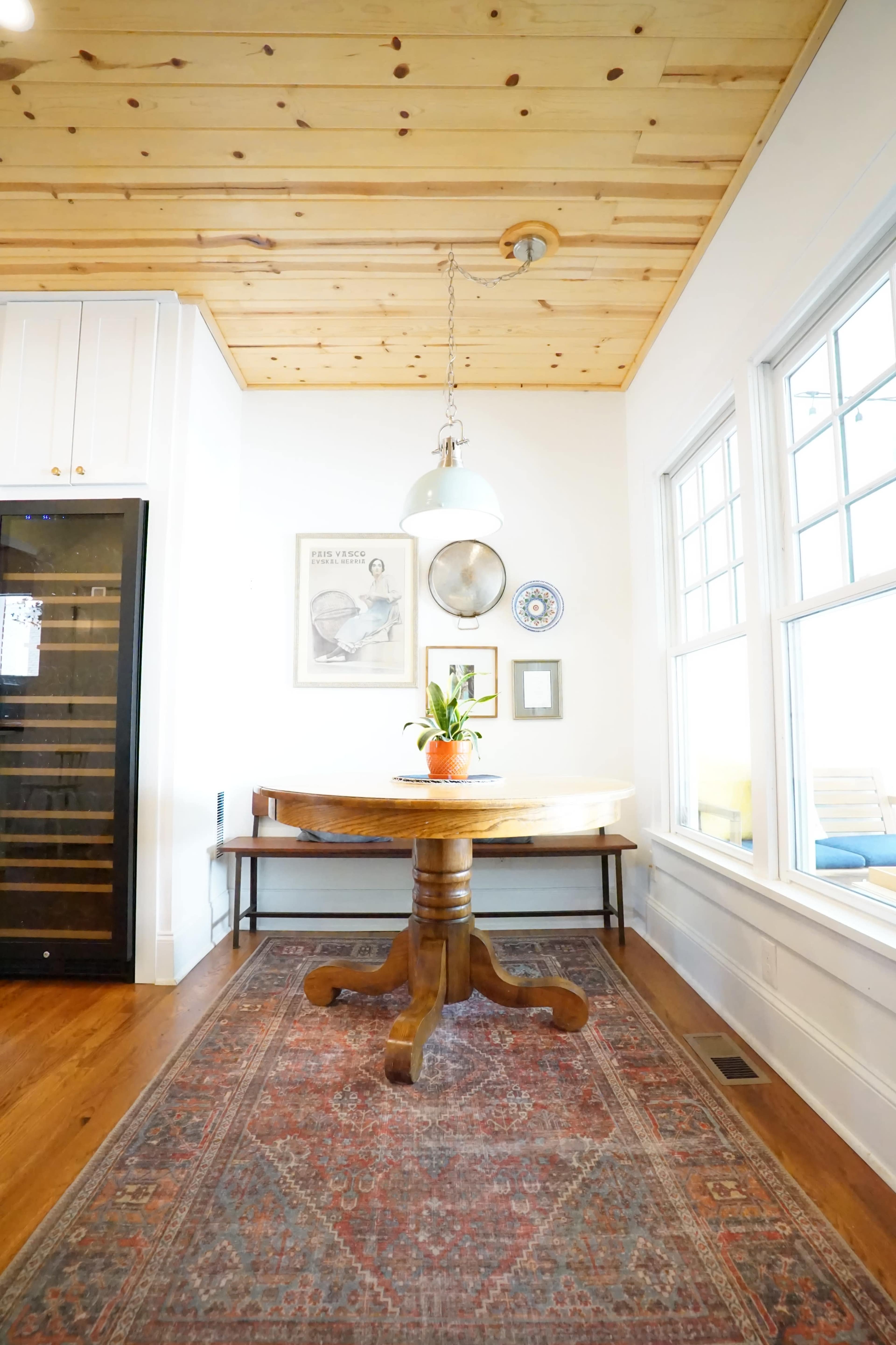 A wooden table with a potted plant sits under a pendant light in a room with a wooden ceiling and large windows.