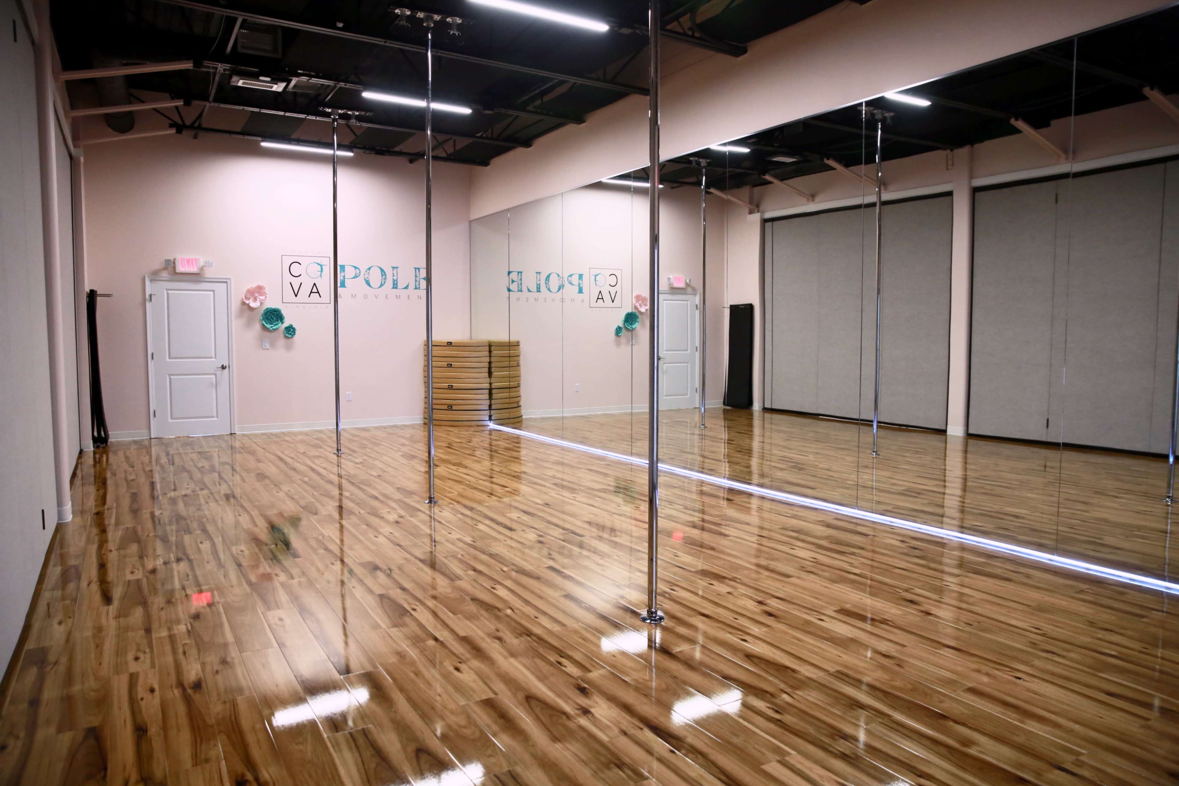 The image shows an empty dance studio with polished wooden floors, two vertical dance poles, and mirrors along the walls.