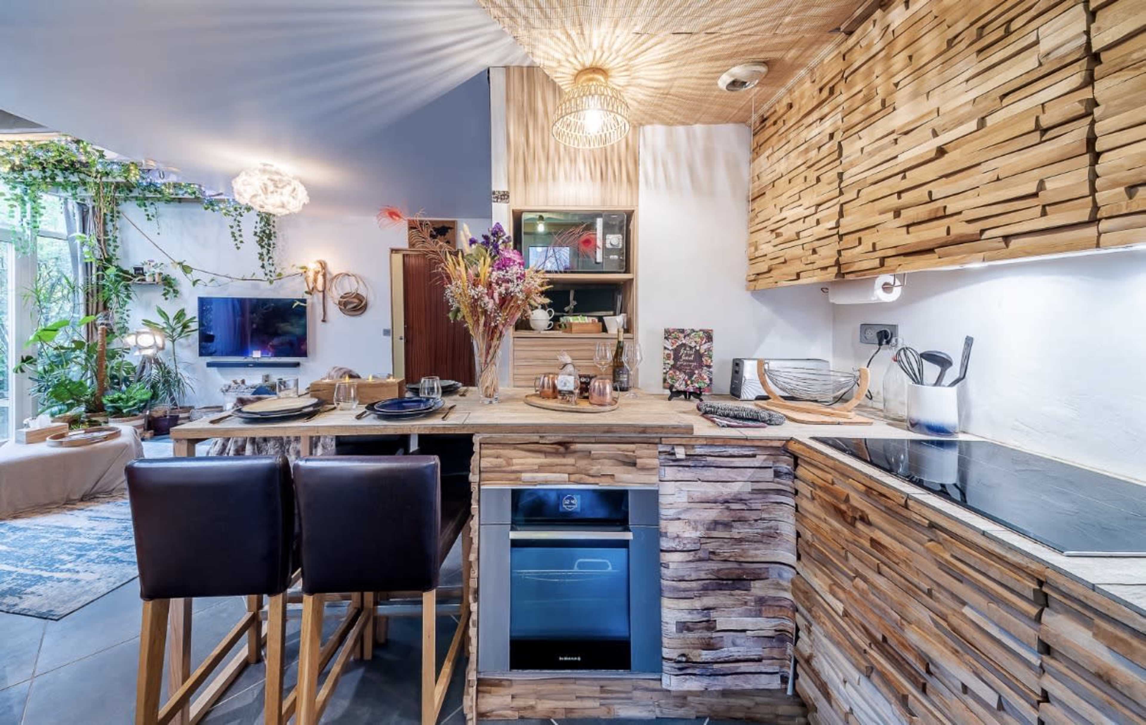 The image shows a modern kitchen with wooden cabinetry, a two-person dining counter, and decorative plants adding greenery to the space.