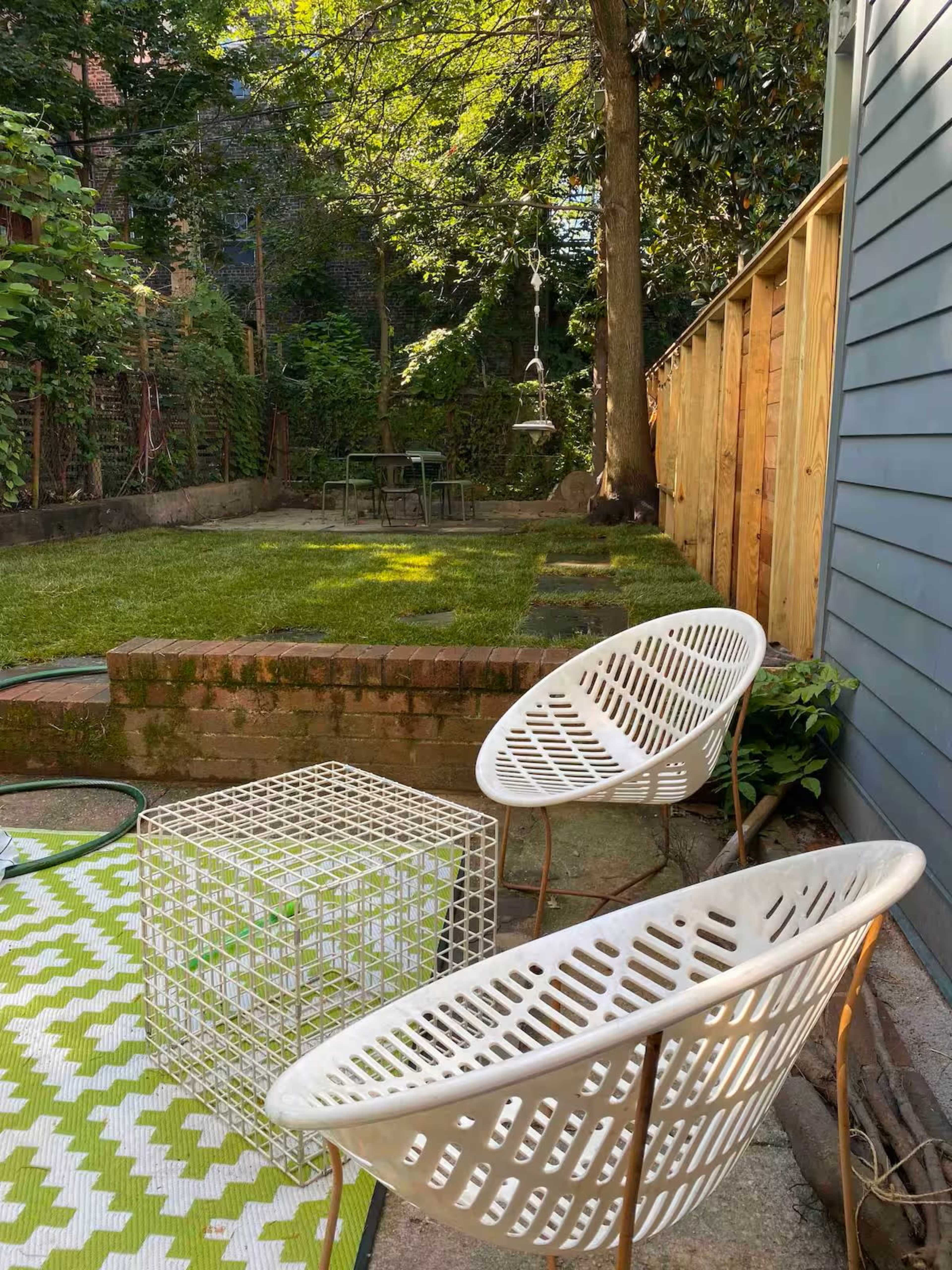 The image shows a tranquil backyard with two white chairs, a geometric table, and a well-maintained lawn surrounded by trees and a wooden fence.