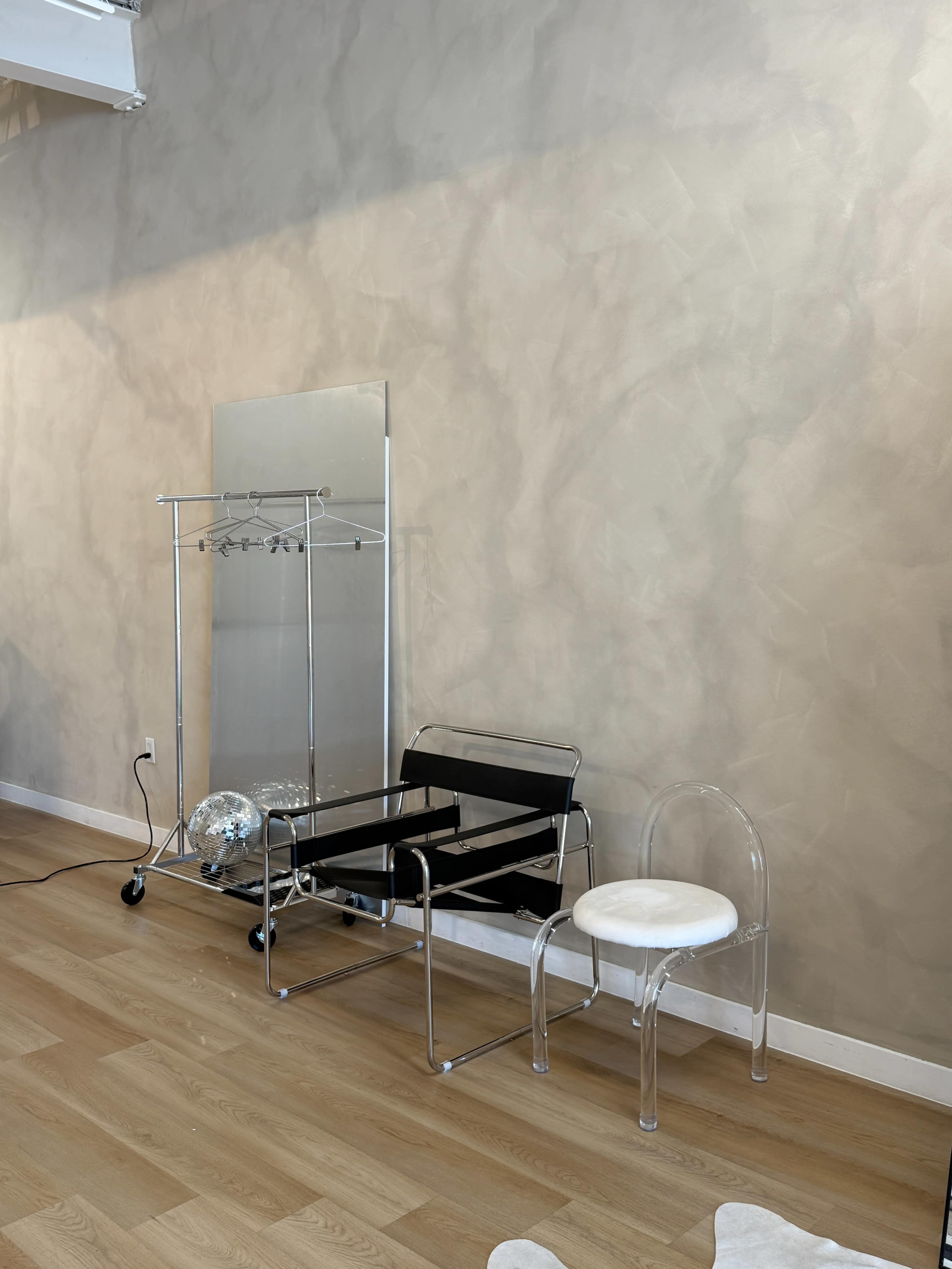 The image displays a minimalist interior featuring a large, textured wall, two chairs—one black and one transparent—alongside a clothing rack and a mirror.
