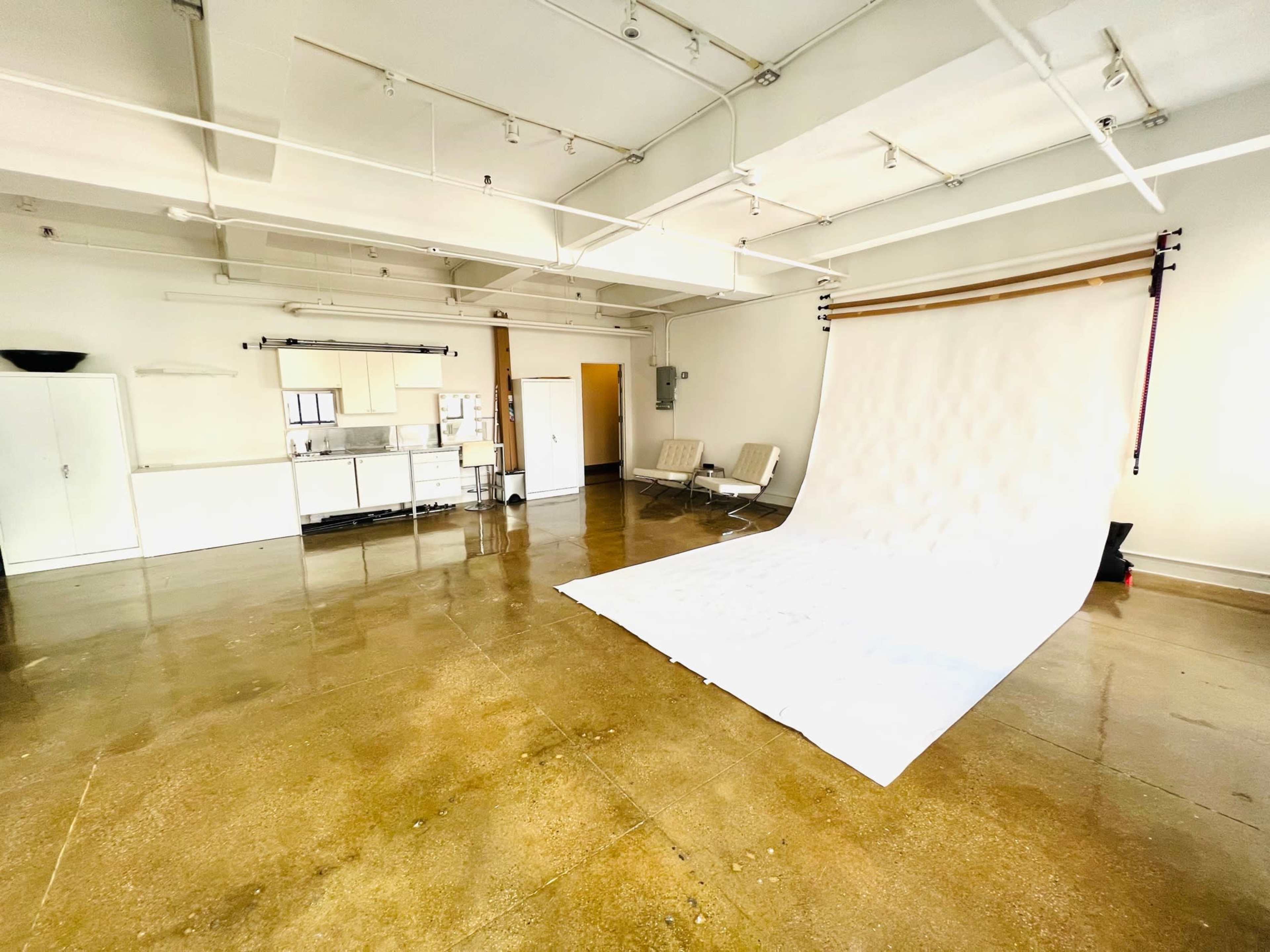 The image shows a spacious photography studio with a white backdrop, two chairs, and a kitchenette area.