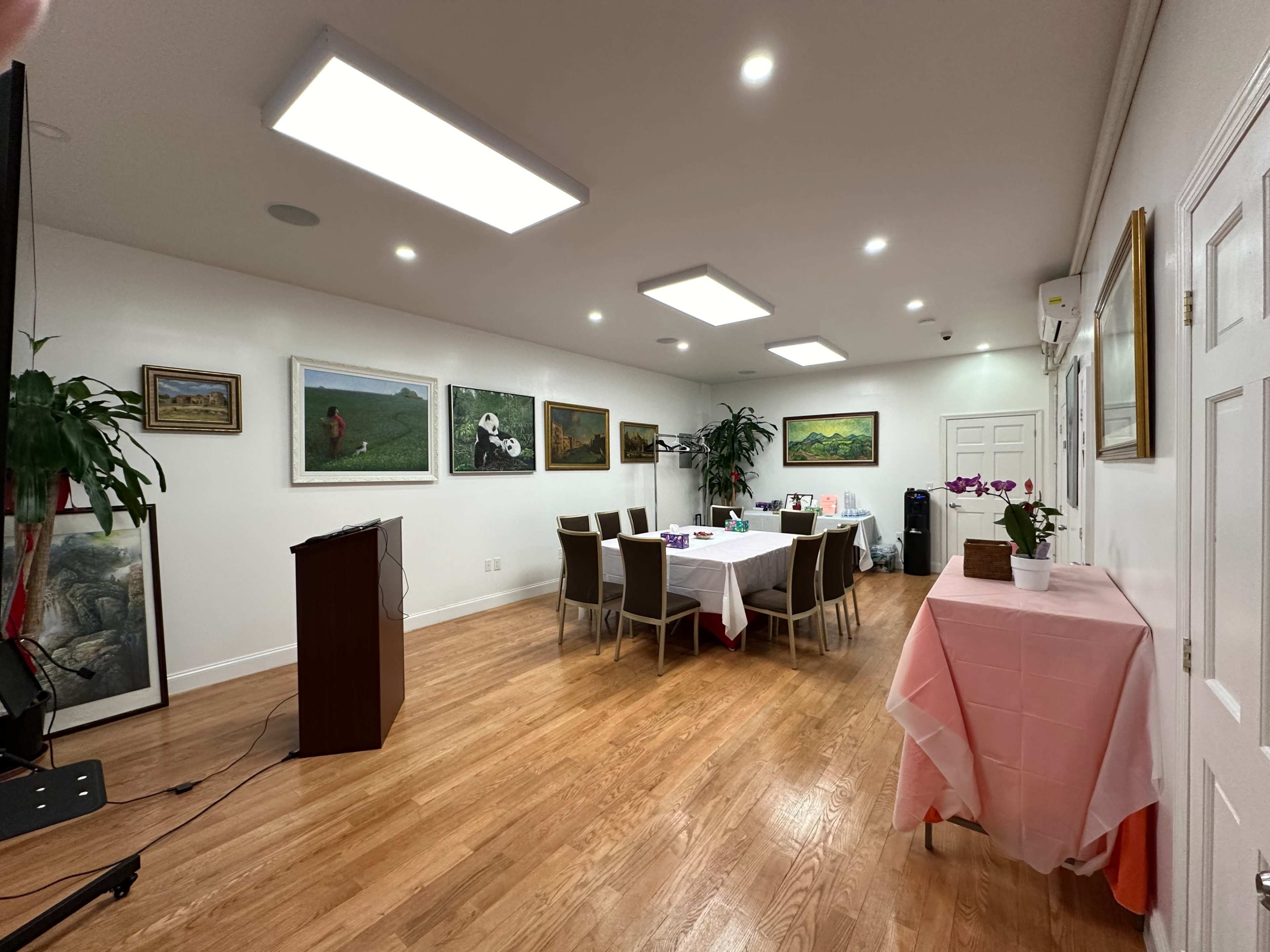 The image shows a well-lit meeting room with a long table set for a gathering, surrounded by chairs and adorned with framed paintings on the walls.