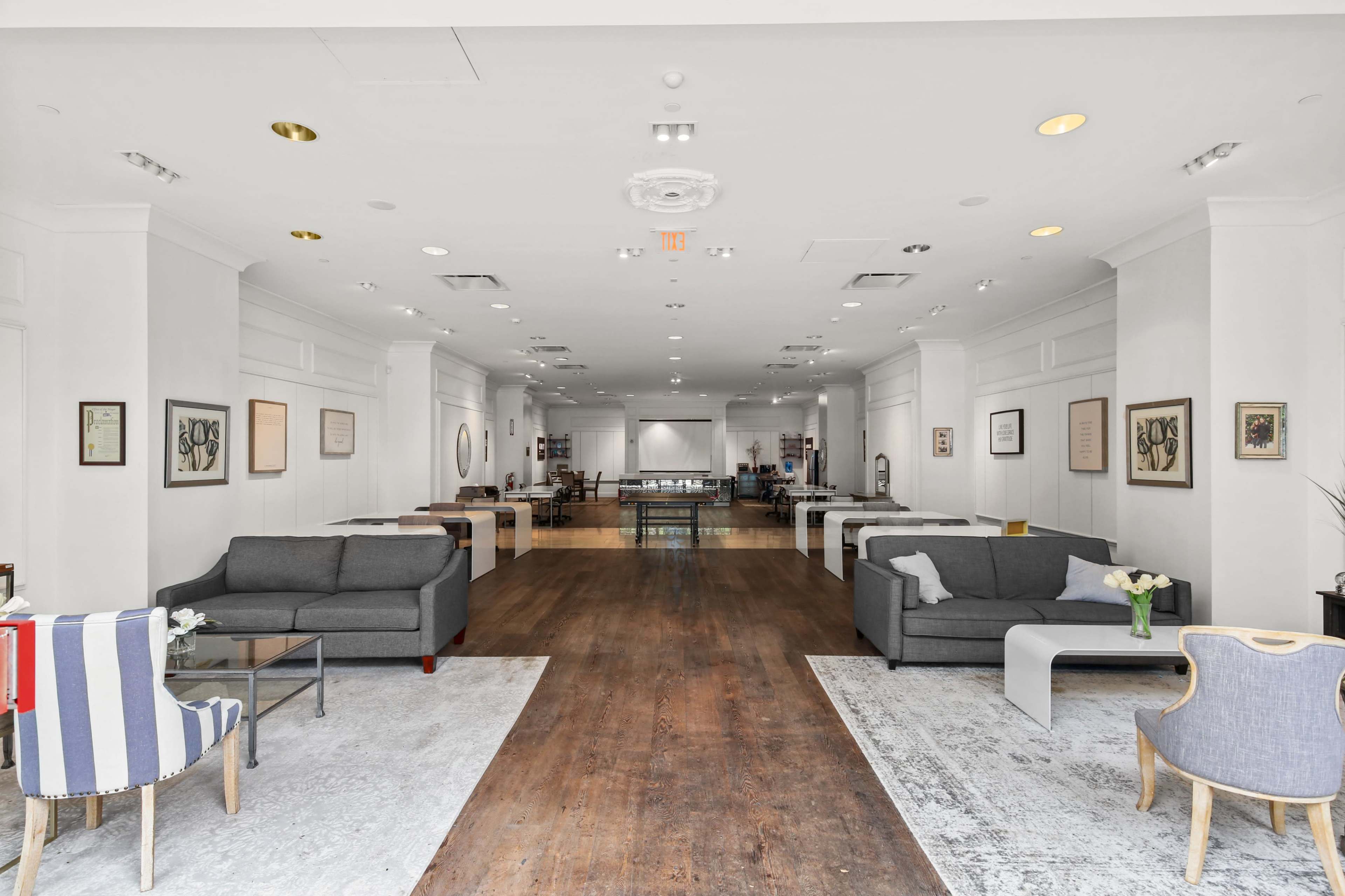 A spacious, well-lit interior features a series of gray sofas and decorative rugs along a wooden floor, with artwork displayed on the walls.