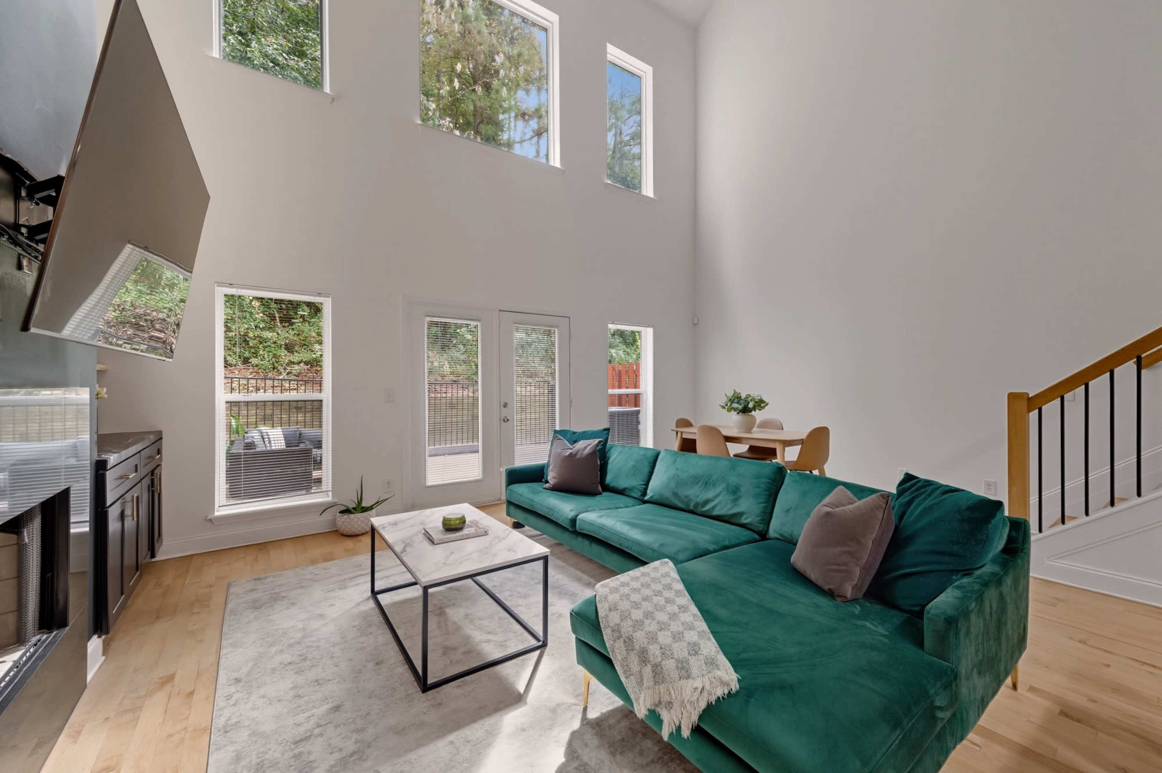The image shows a bright, modern living room with large windows, a green sectional sofa, a coffee table, and a dining area visible in the background.