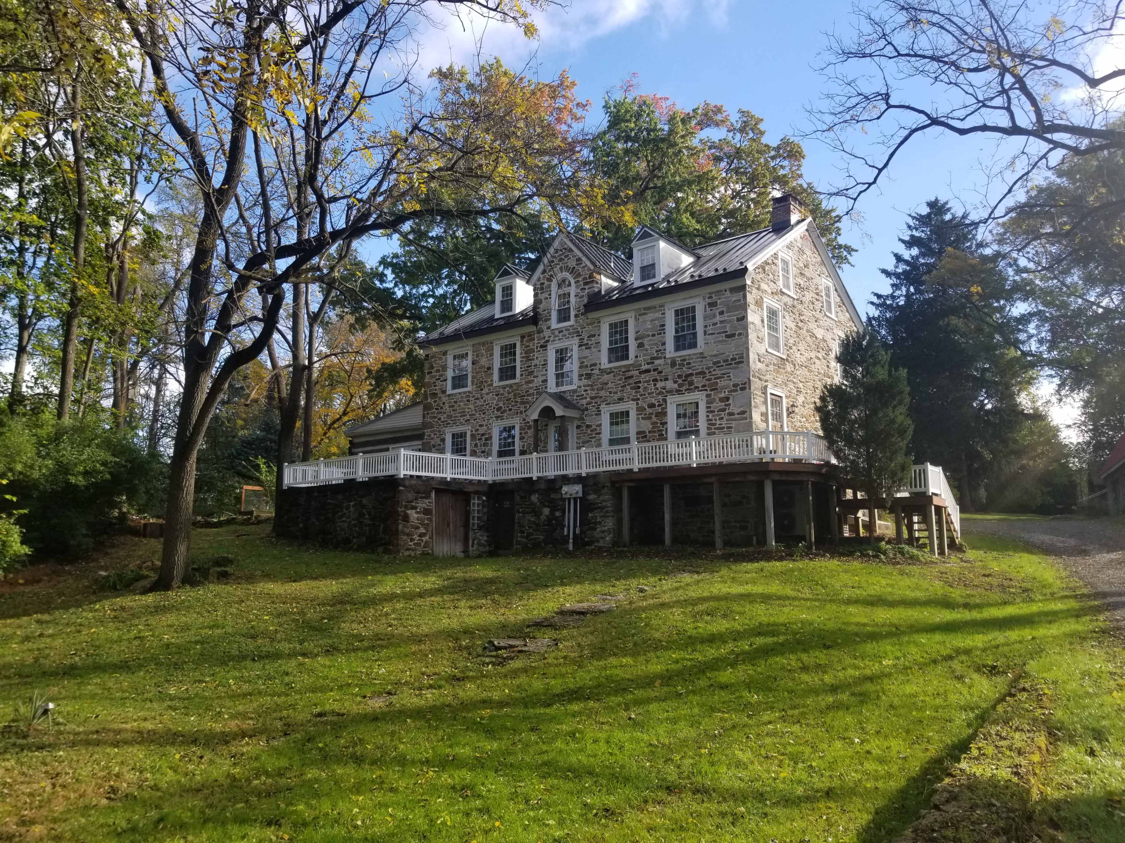 A large stone house with multiple gables and a deck, situated on a hillside surrounded by trees and a grassy area.