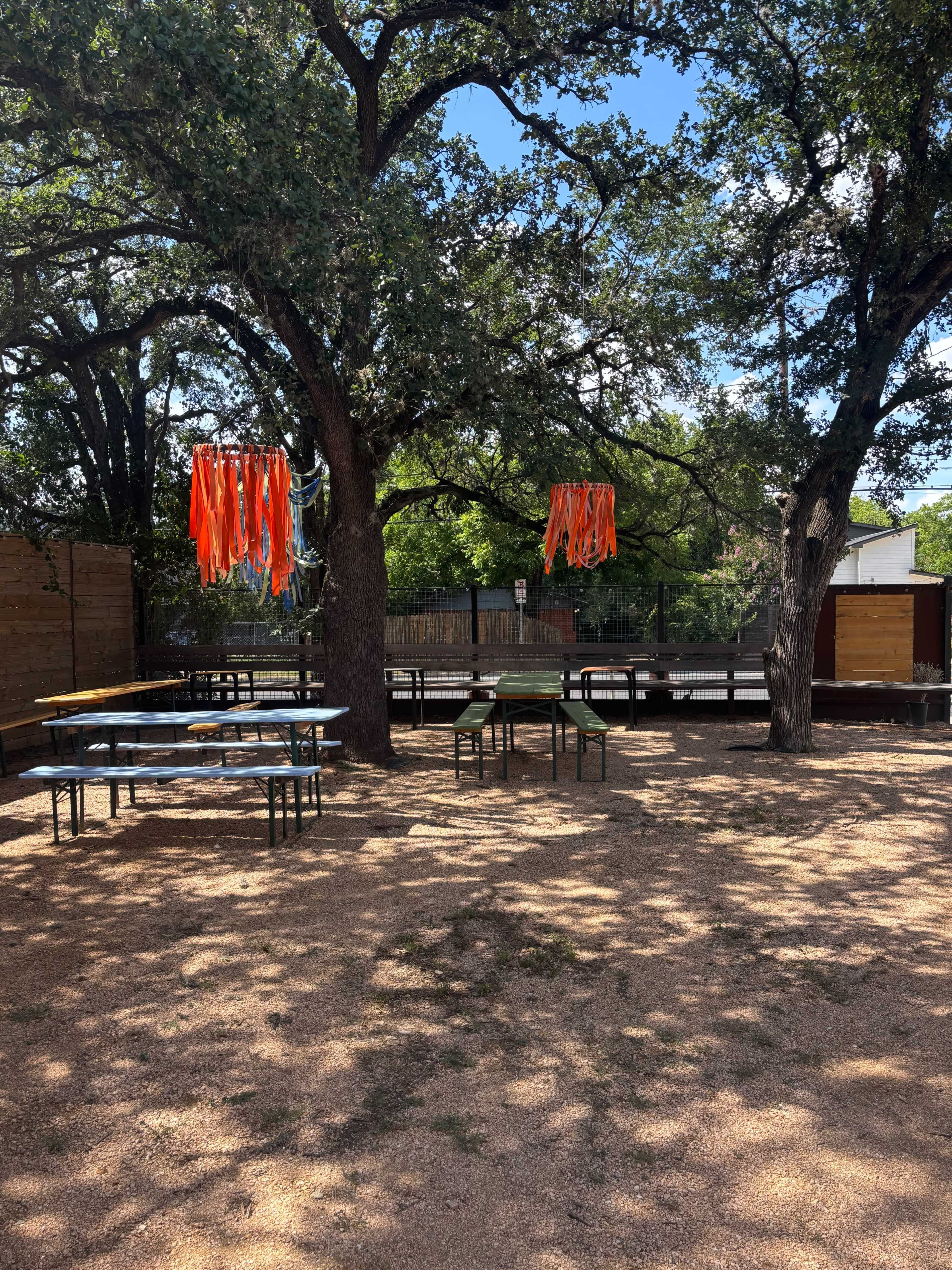 The image shows a gravel area with two trees hanging with bright orange ribbons, surrounded by tables and a fenced background.