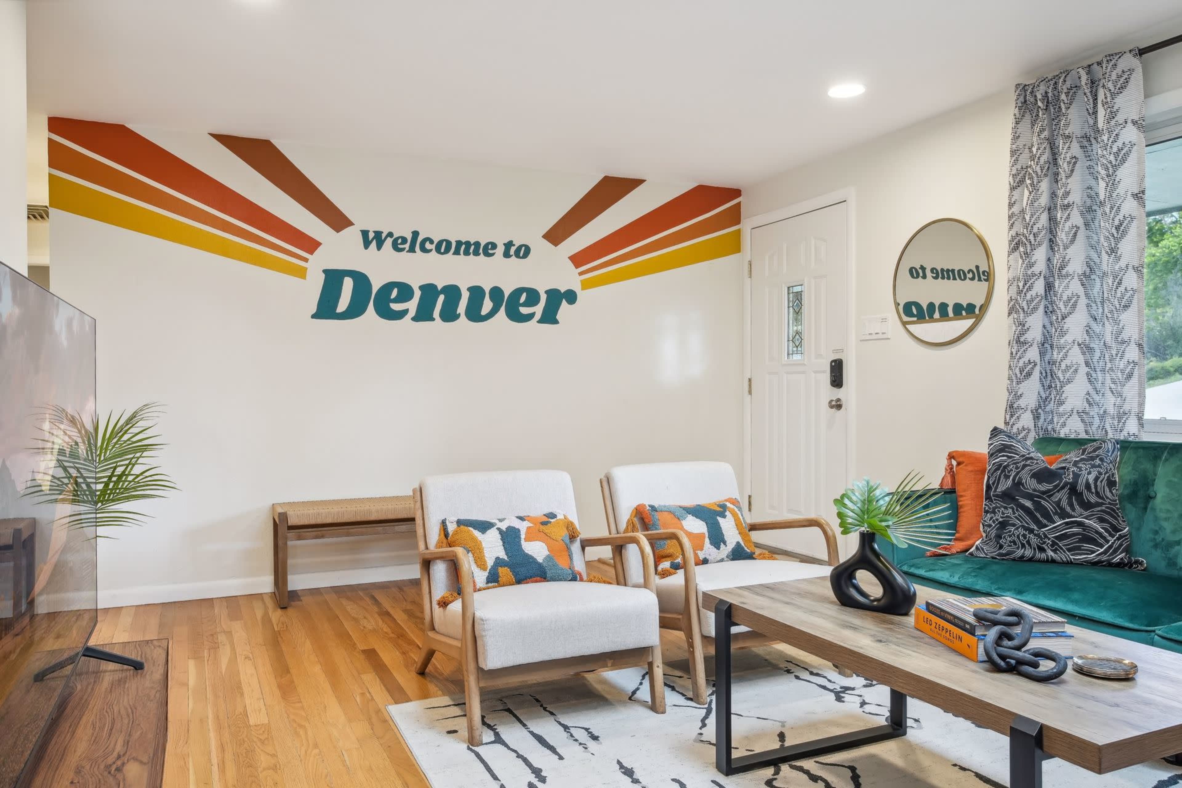 The living room features a welcoming mural that reads "Welcome to Denver," paired with modern furniture and natural wooden accents.