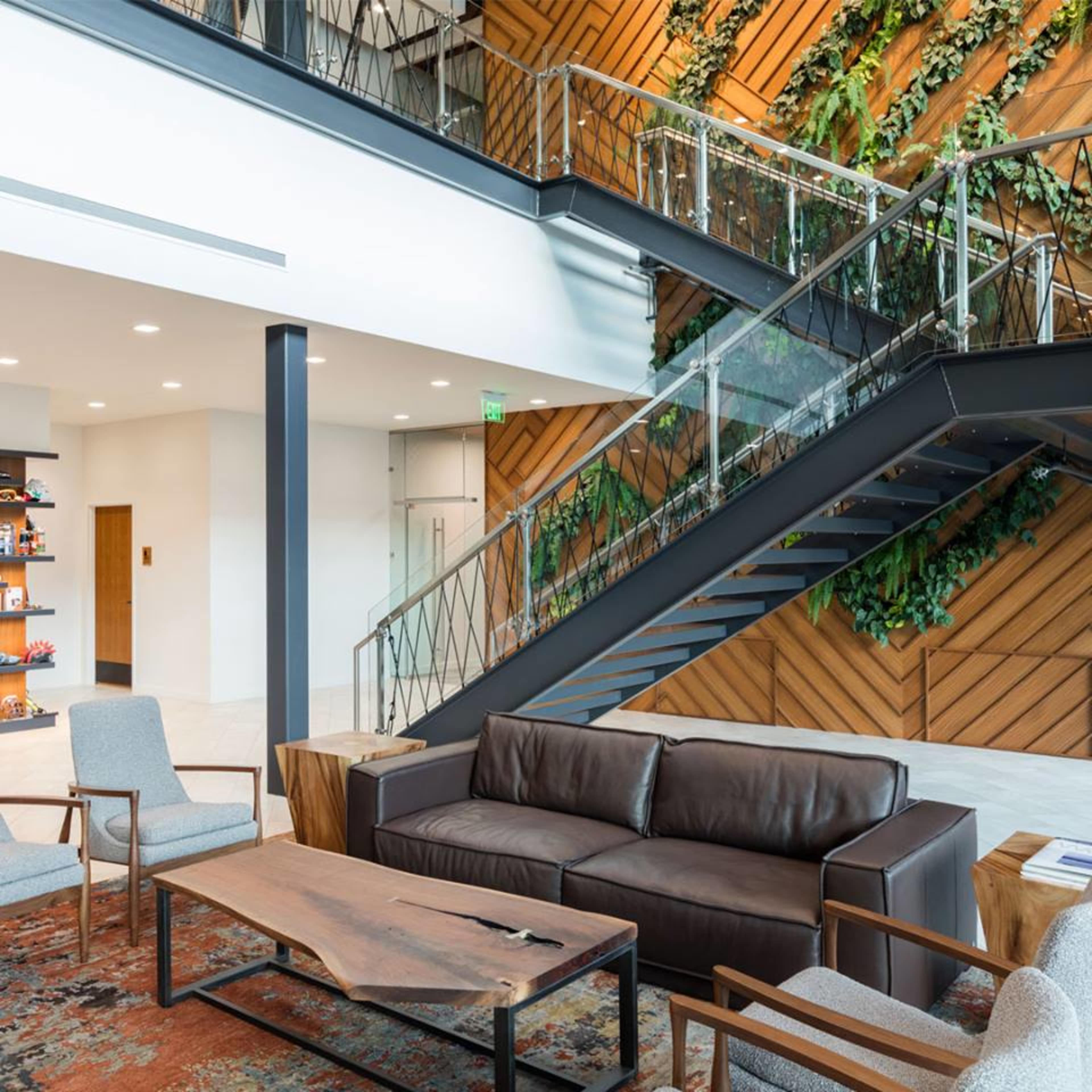 The image shows a modern interior space featuring a staircase with glass railings, wooden walls adorned with plants, and a seating area with a brown leather sofa and wooden tables.