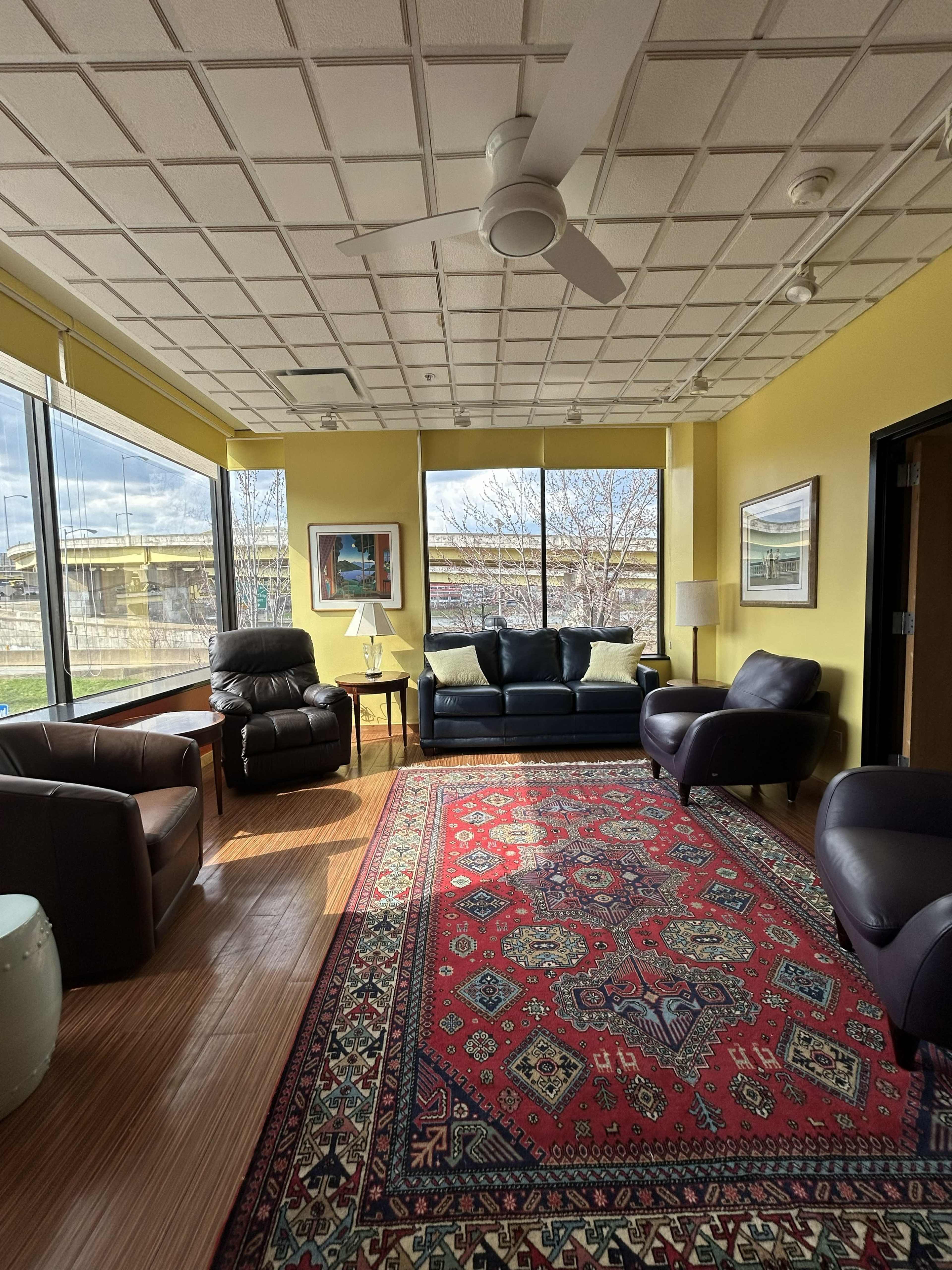 The image shows a cozy living room with large windows, a dark blue couch, two armchairs, and a patterned rug on wooden flooring.