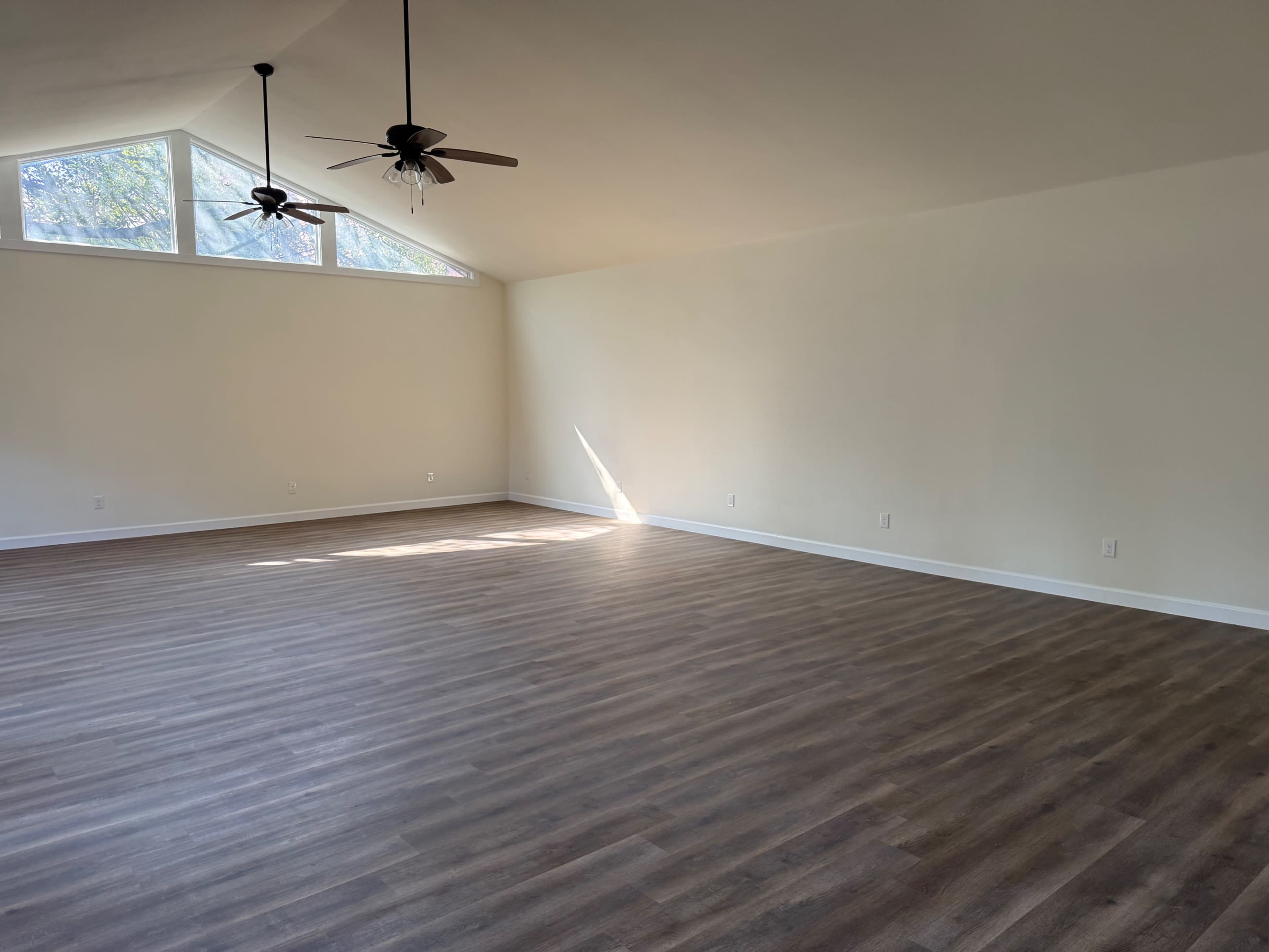 An empty room with high ceilings, two ceiling fans, and large windows allowing natural light to enter.