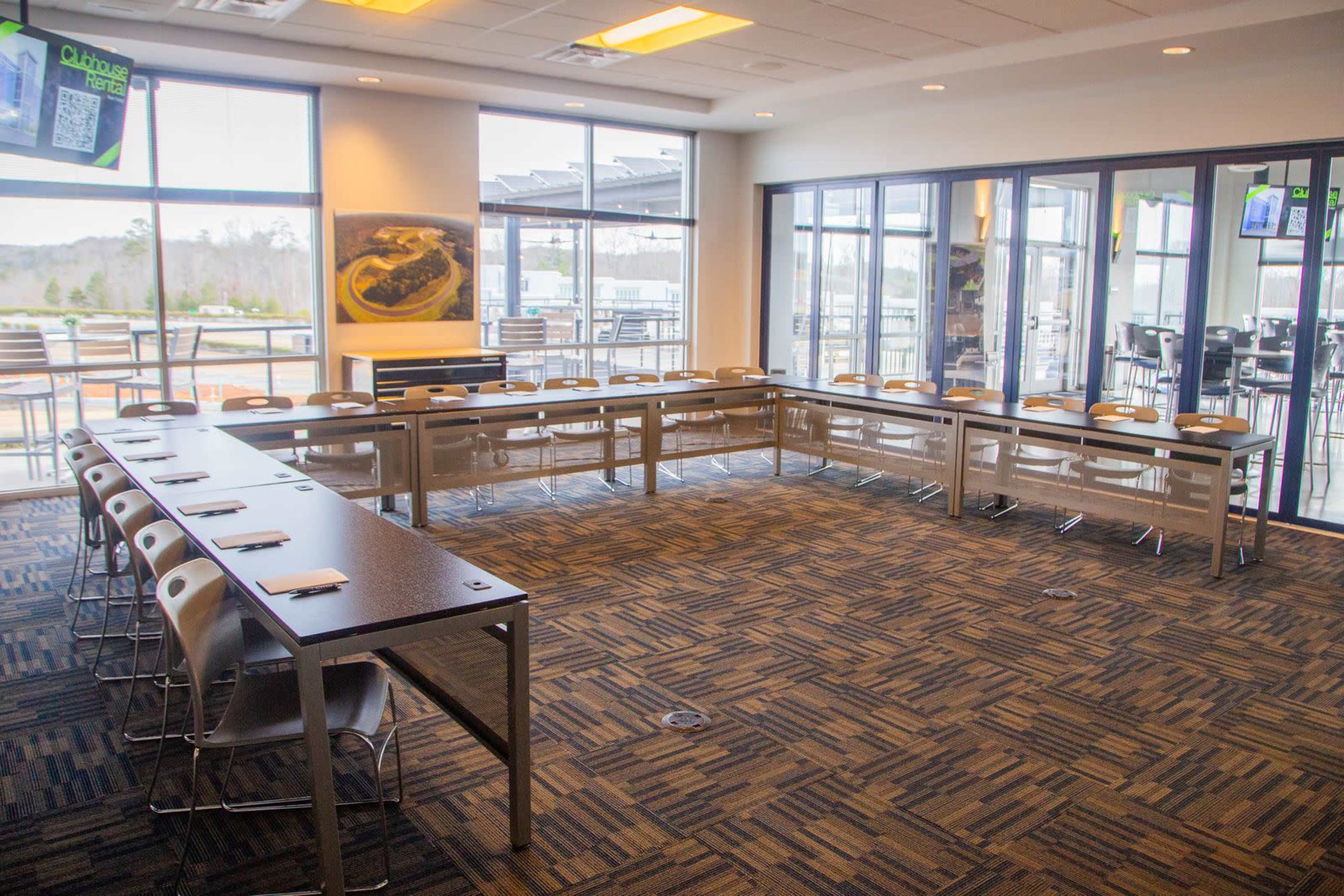 The image shows a spacious meeting room arranged with multiple tables and chairs in a circular layout, featuring large windows that provide natural light and a view of an outdoor area.