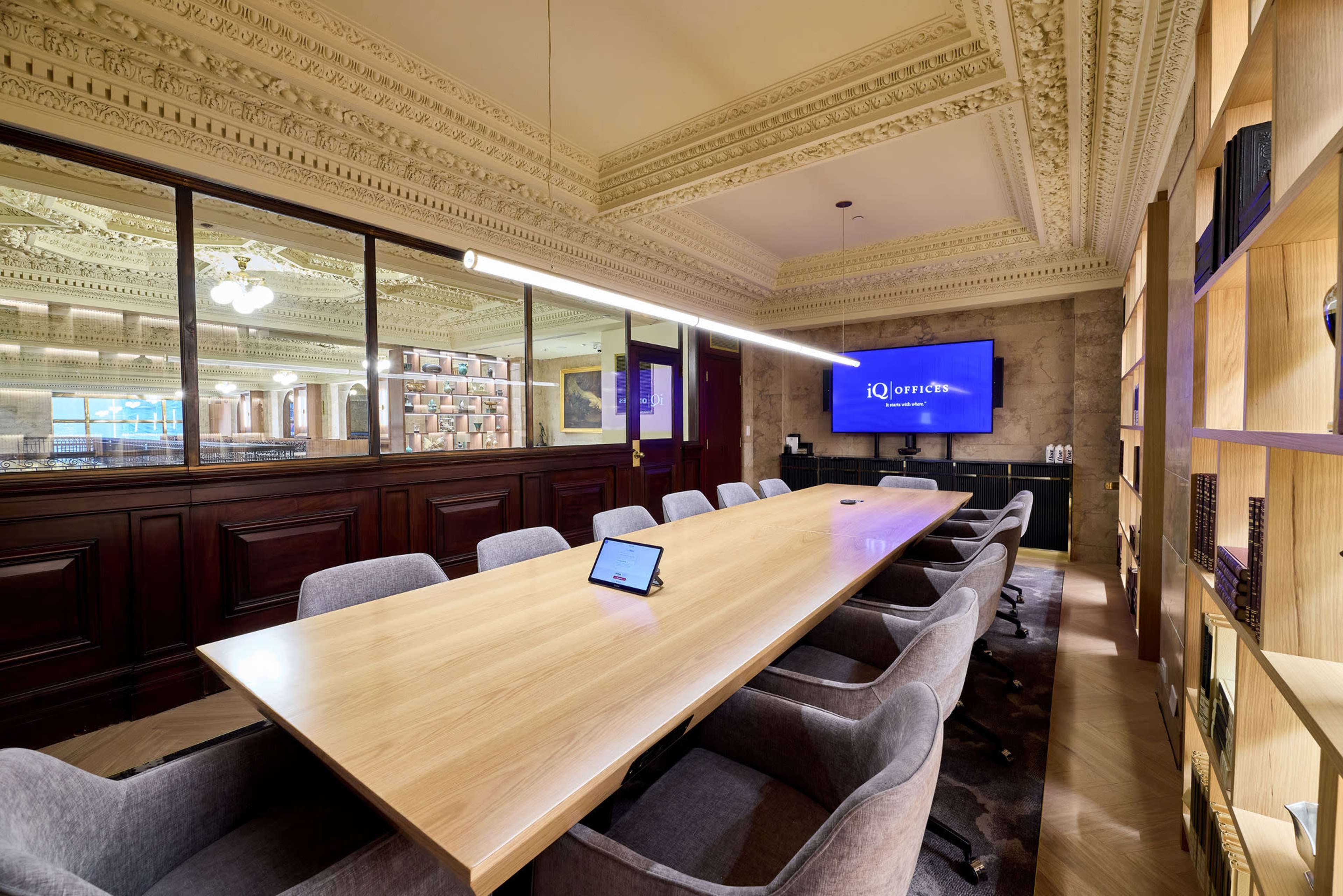 A long wooden conference table with upholstered chairs is set in a modern meeting room featuring large windows and a wall-mounted screen.