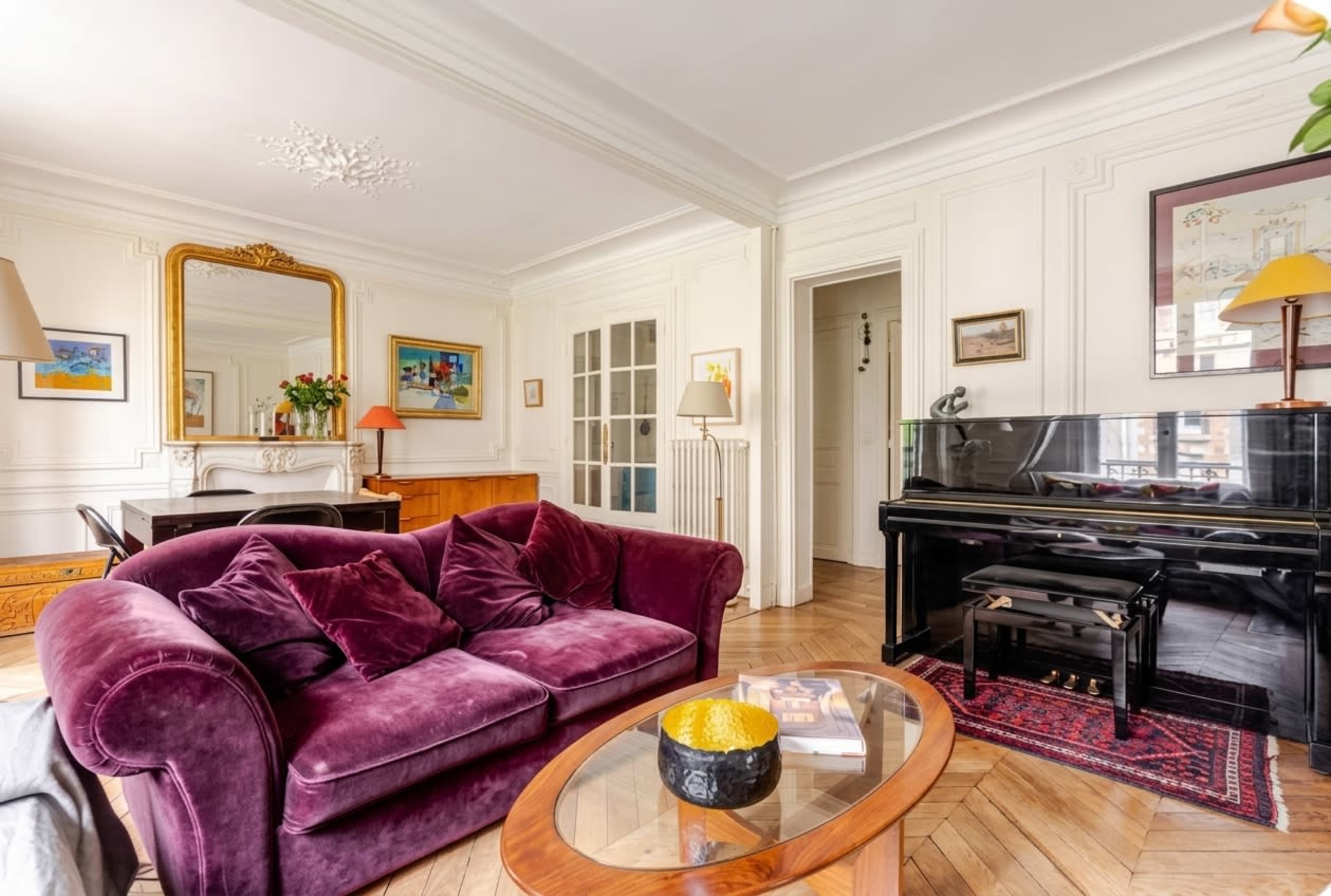 The image shows a cozy living room featuring a purple sofa, a glass coffee table, and a black piano against a backdrop of white walls and wooden flooring.