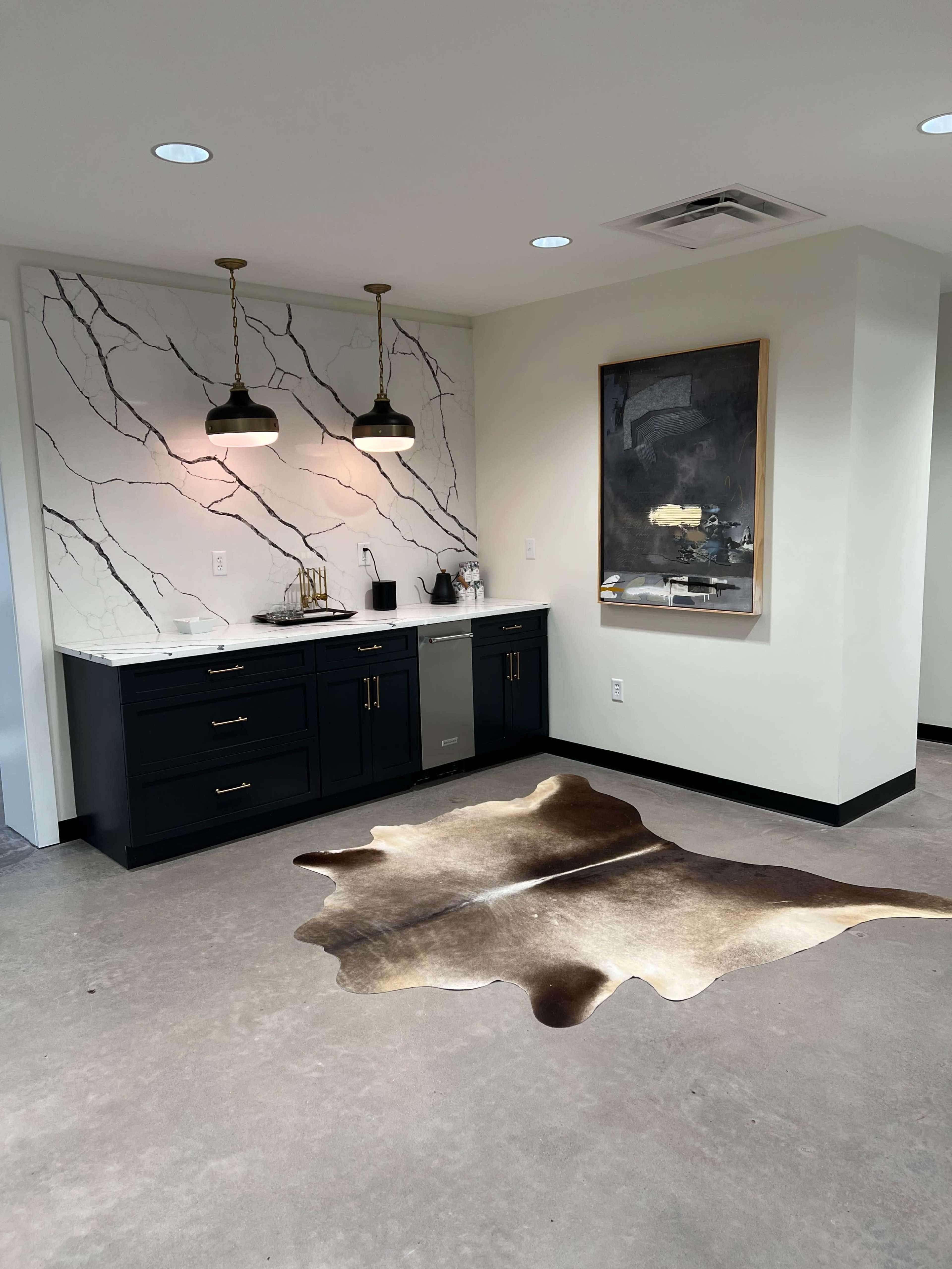 A modern kitchen area with dark cabinetry, a marble-patterned wall, pendant lighting, and a cowhide rug on the concrete floor.