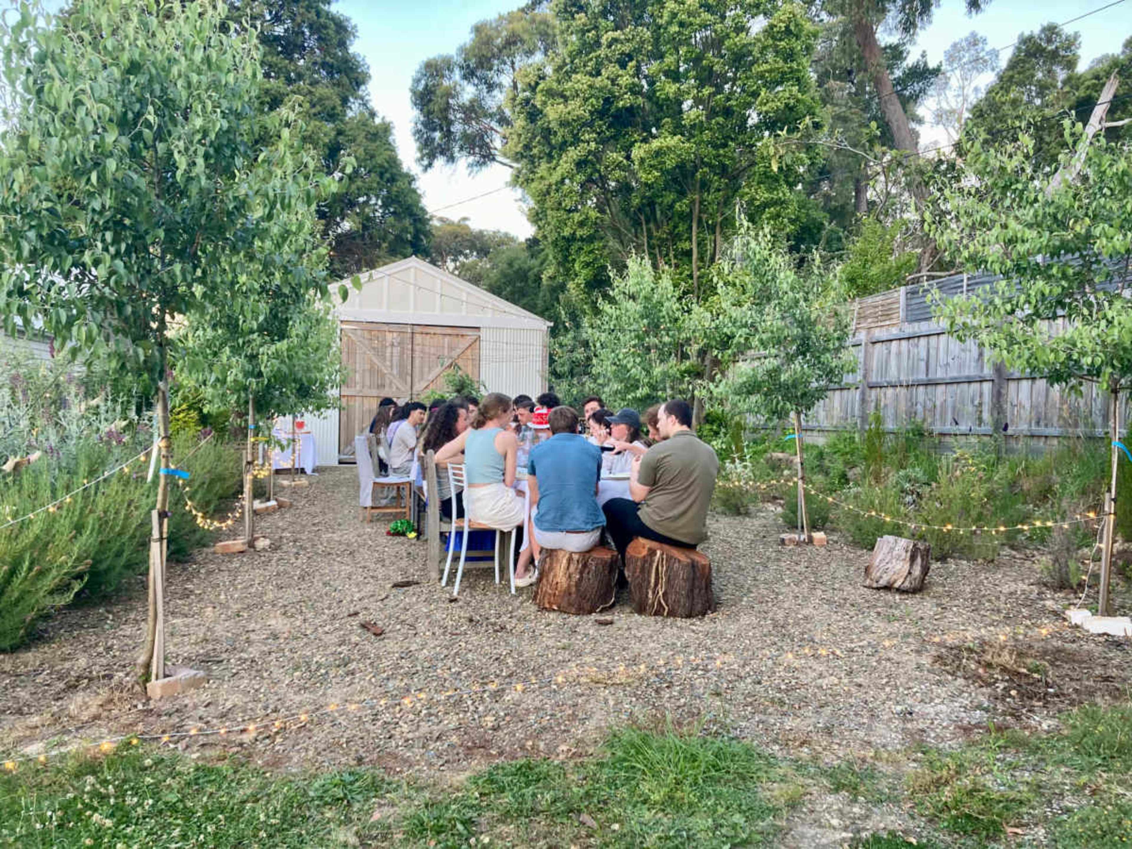 A group of people sits at a long table outdoors in a garden surrounded by trees and a greenhouse.