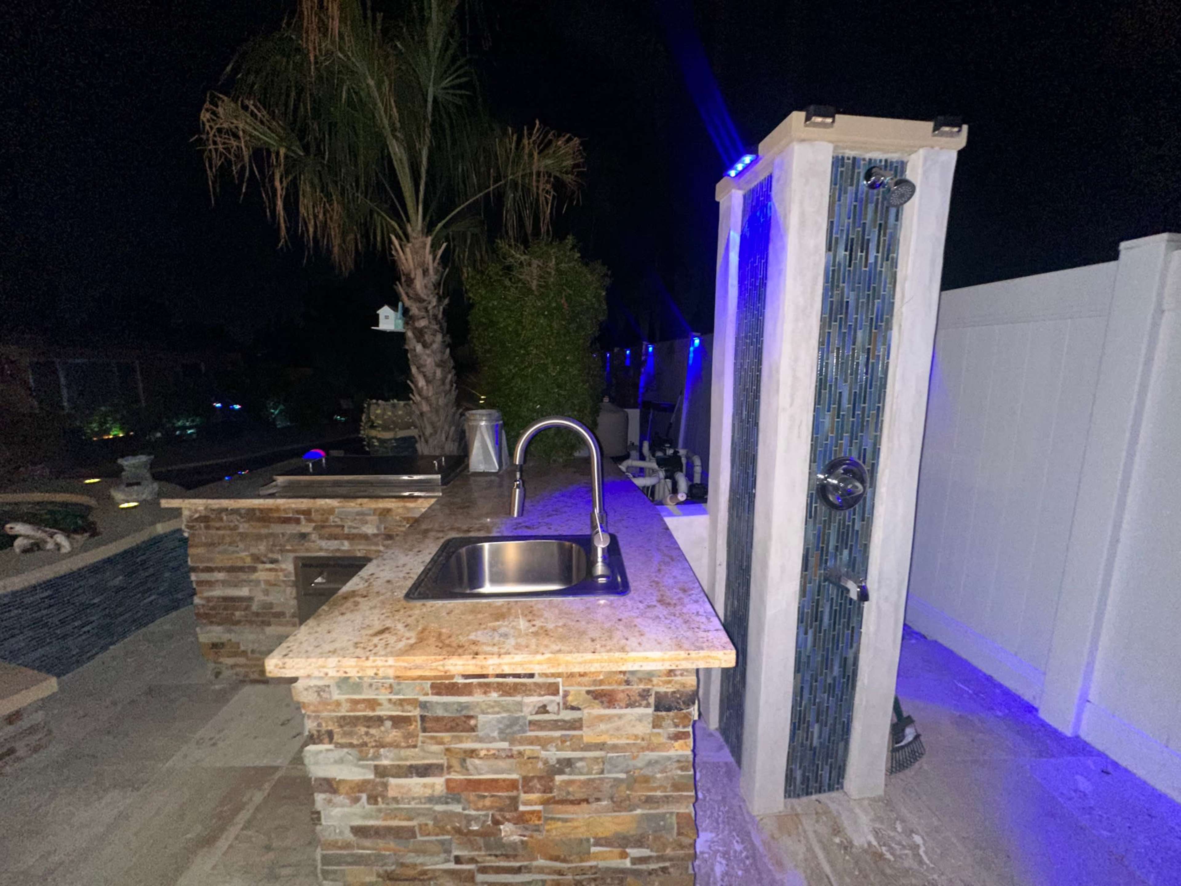 The image shows an outdoor kitchen area at night, featuring a stone countertop with a sink and an adjacent shower, illuminated by blue lights and surrounded by palm trees.