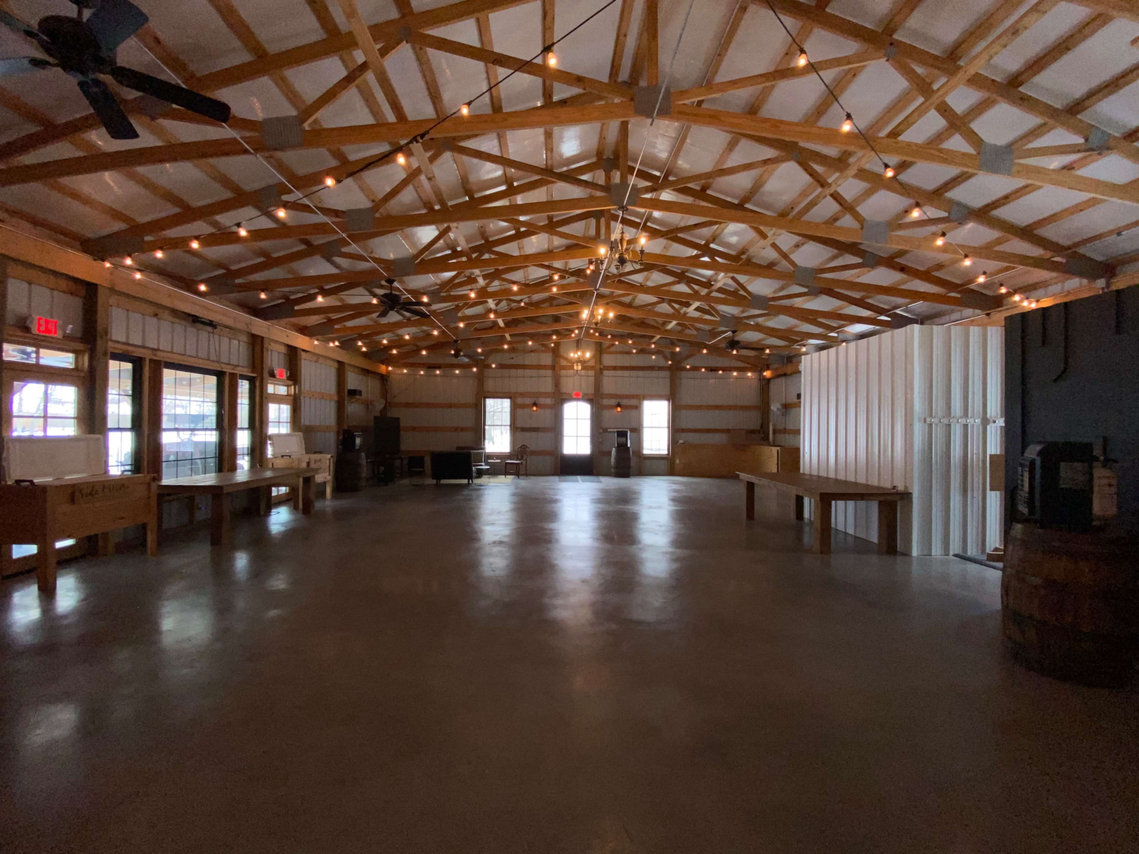 The image shows an empty indoor venue with wooden beams, string lights overhead, and a polished concrete floor.